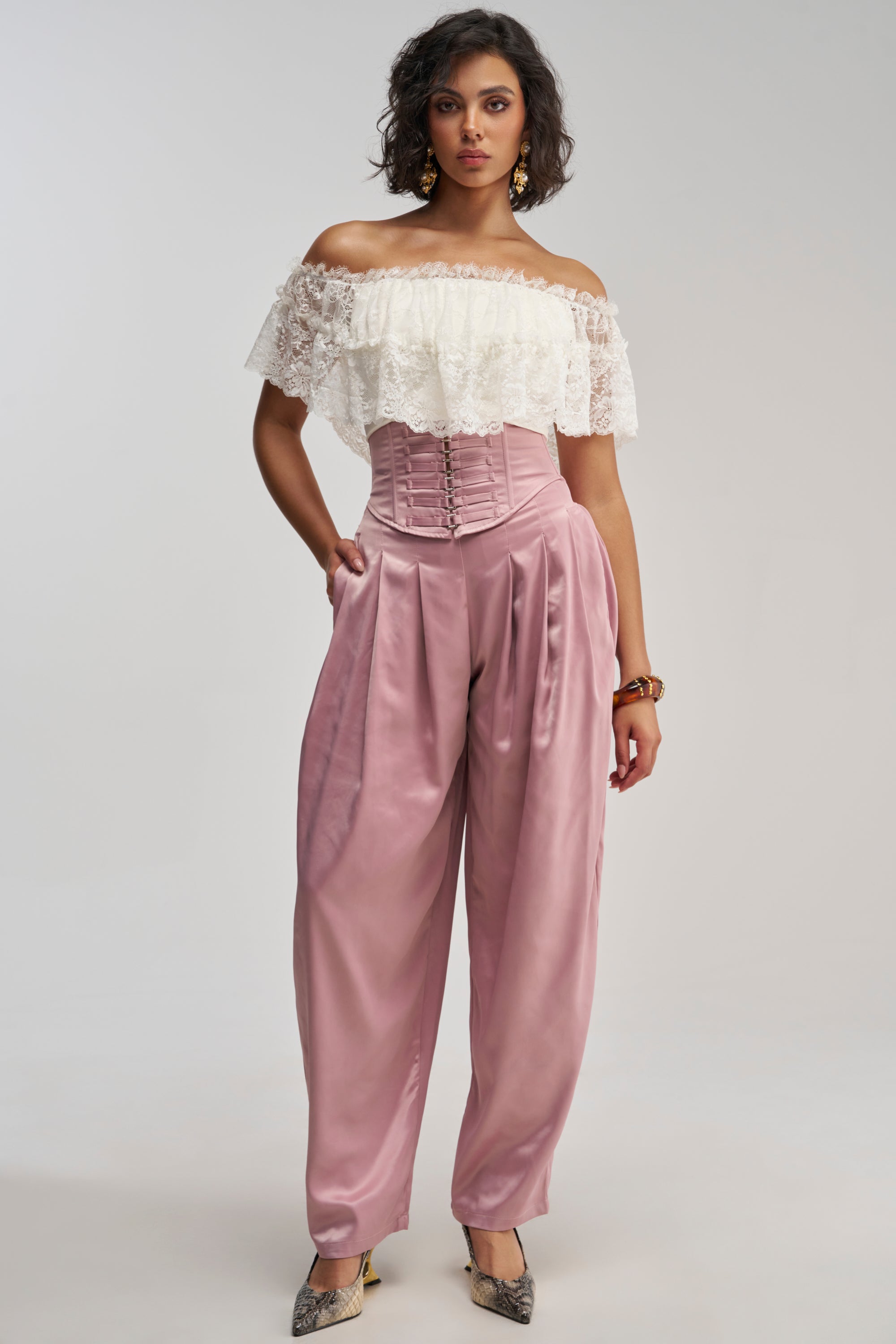 Wearing the DRESS LIKE ME CORSETED TROUSER, a woman poses in high-waisted, mauve satin pants with a corset-like waistband. She pairs them with a white off-shoulder lace top, accessorized with earrings, a bracelet, and pointed shoes as she stands against a plain background.