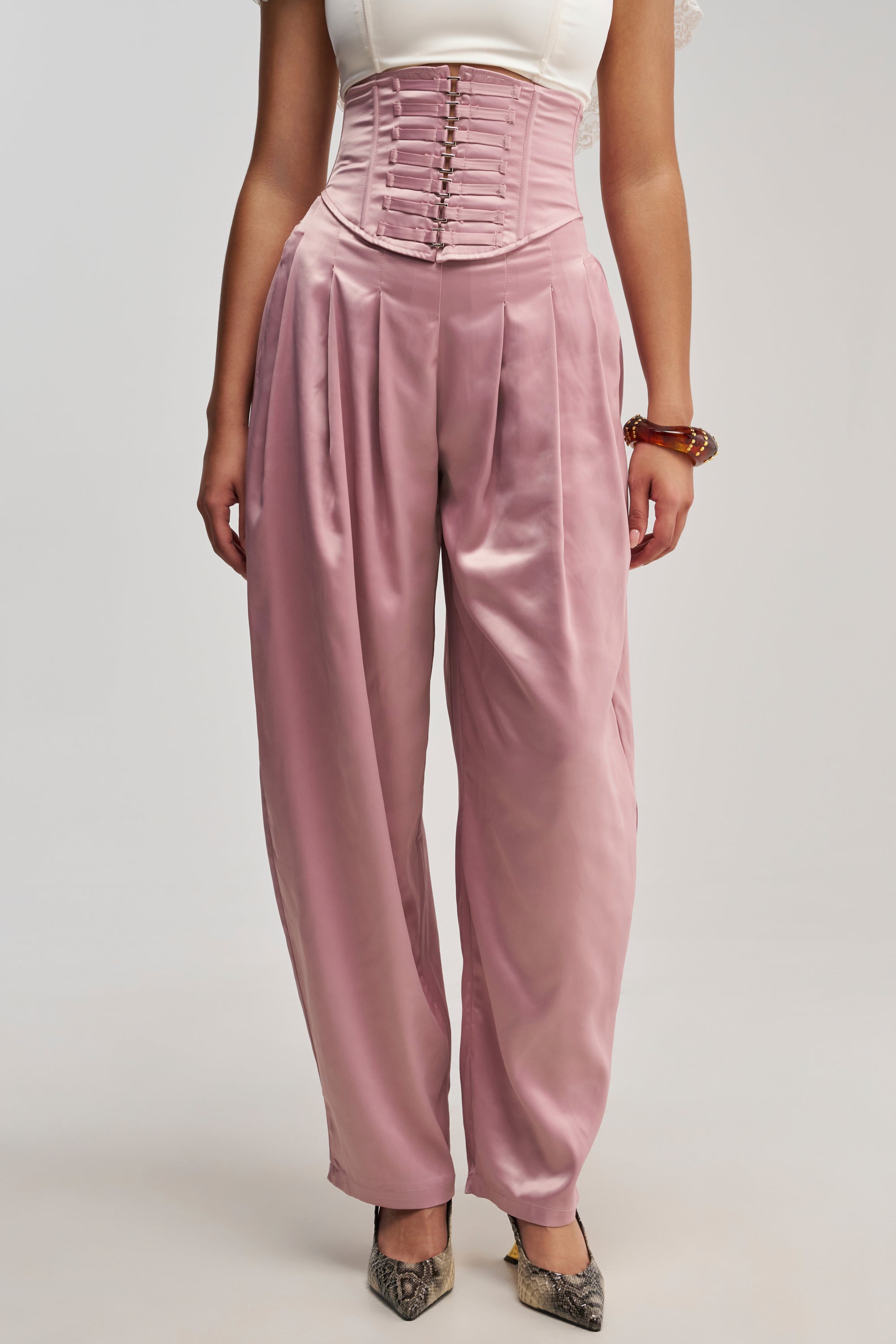 Wearing the DRESS LIKE ME CORSETED TROUSER—high-waisted, mauve satin trousers featuring pleats and a corset waist—a woman pairs them with a white top, a chunky bracelet, and snakeskin-patterned shoes to create a chic balloon silhouette.