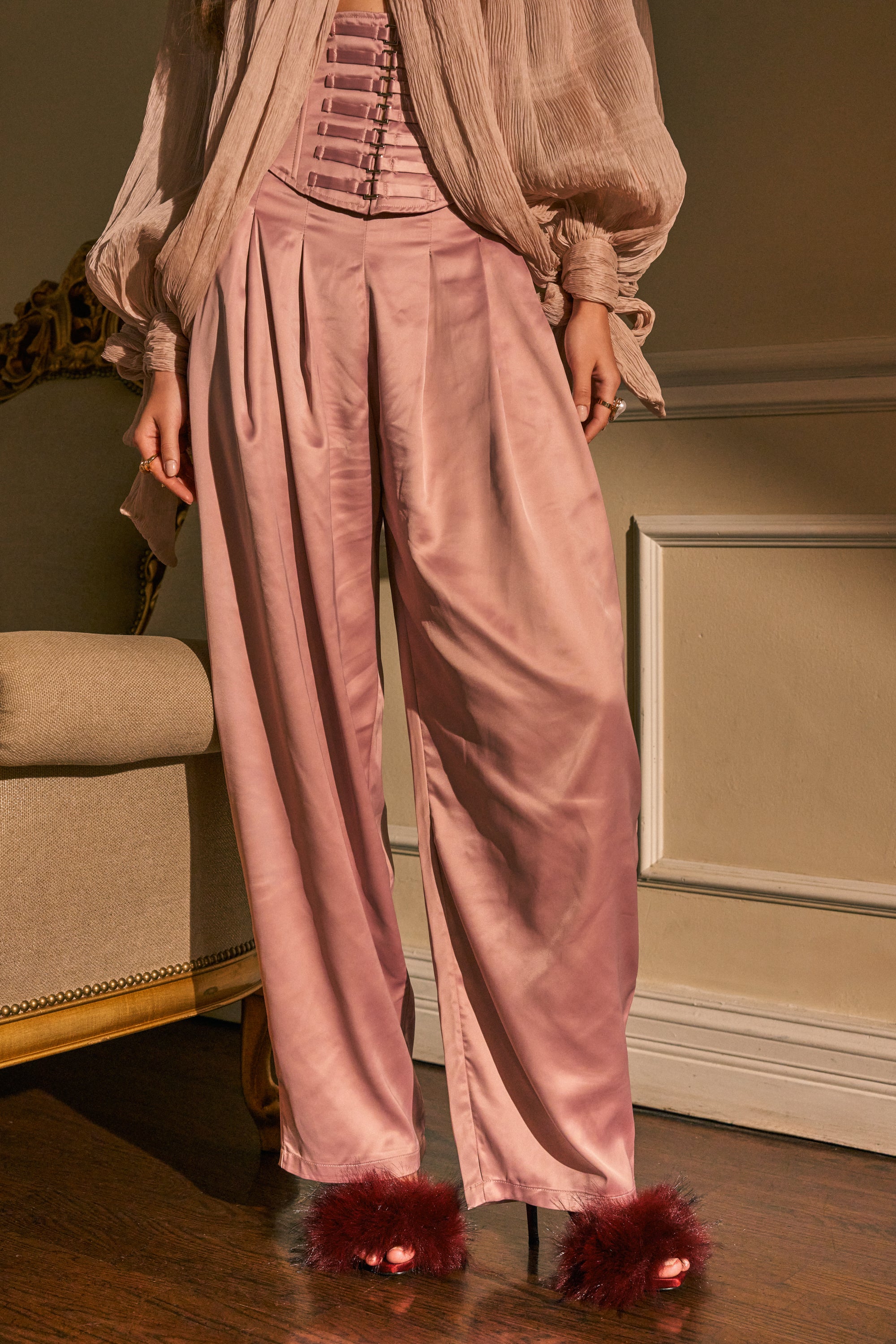 Wearing the DRESS LIKE ME CORSETED TROUSER—a pair of loose, silky, blush-pink pleated pants in a chic balloon silhouette—a person stands indoors on a wooden floor next to a beige chair and a cream wall with molding. They complete the look with a matching corset top, sheer blouse, and fluffy maroon heels.