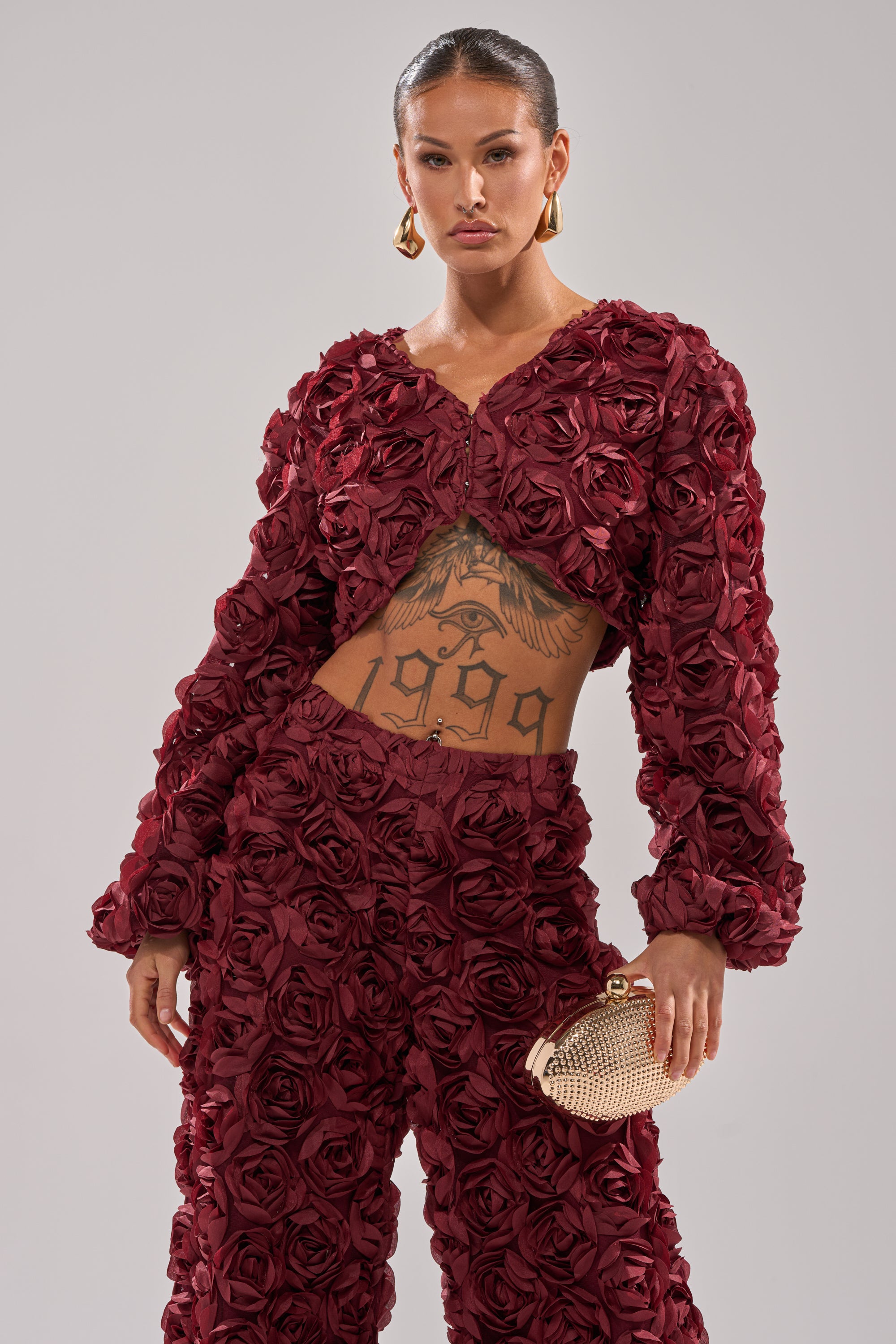 A woman wears the ROSE PETALS FLORAL SHRUG—a burgundy cropped outfit with 3D roses—paired with gold hoop earrings, a gold textured clutch, slicked-back hair, and a striking “1999” tattoo across her chest and stomach. 0
