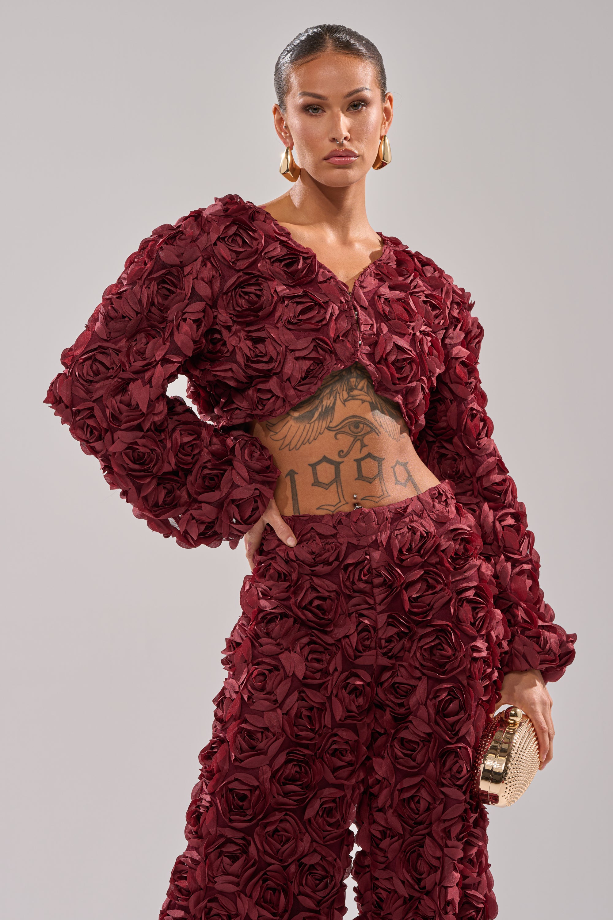 Wearing the ROSE PETALS FLORAL SHRUG in deep red, adorned with 3D fabric roses, this individual pairs it with gold hoop earrings, a sleek hairstyle, a small gold clutch, and reveals an abdominal tattoo. 1