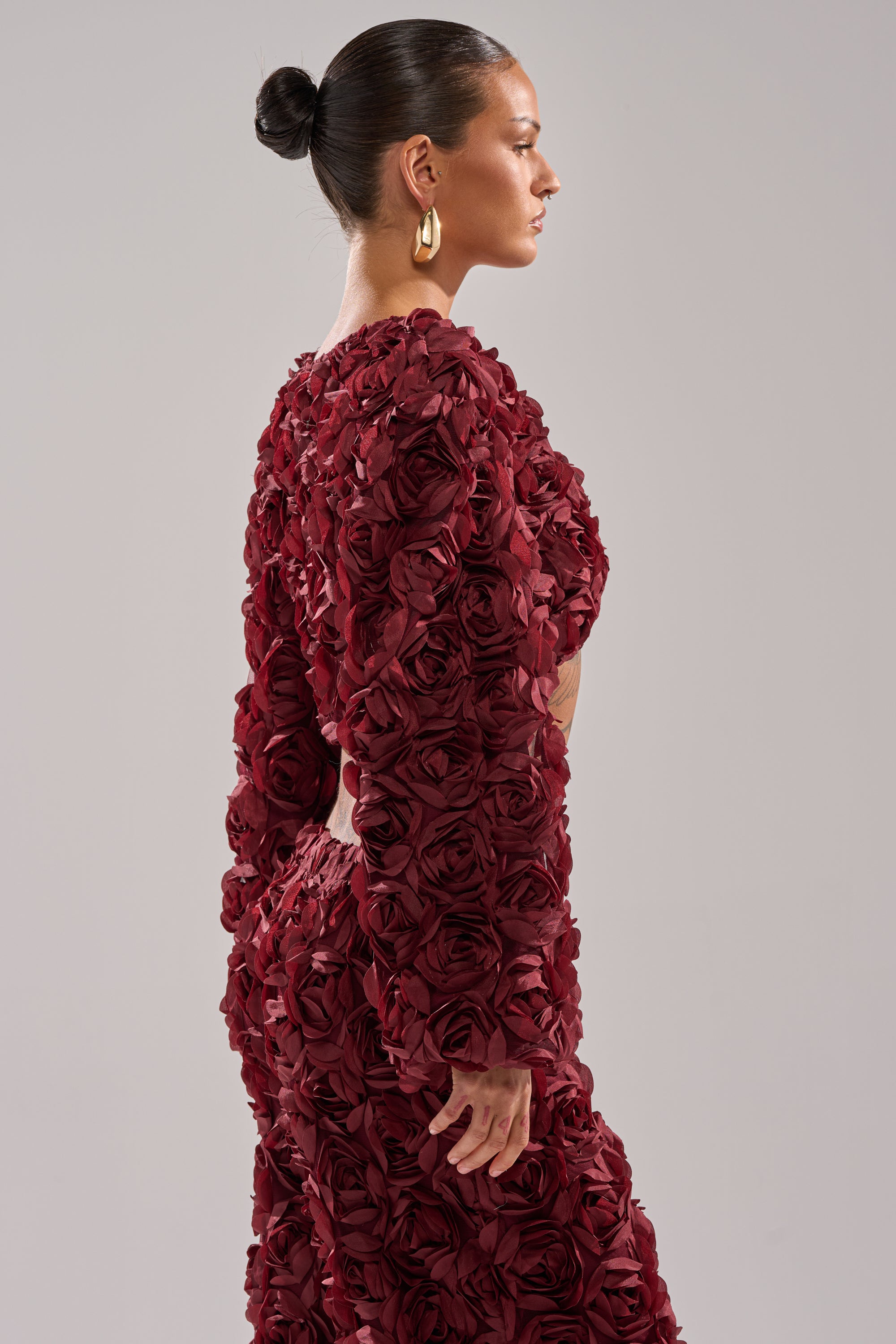 A woman with slicked-back hair wears large gold hoops and the ROSE PETALS FLORAL SHRUG, a textured burgundy cropped jacket adorned with 3D fabric roses, standing in profile against a plain light background. 2