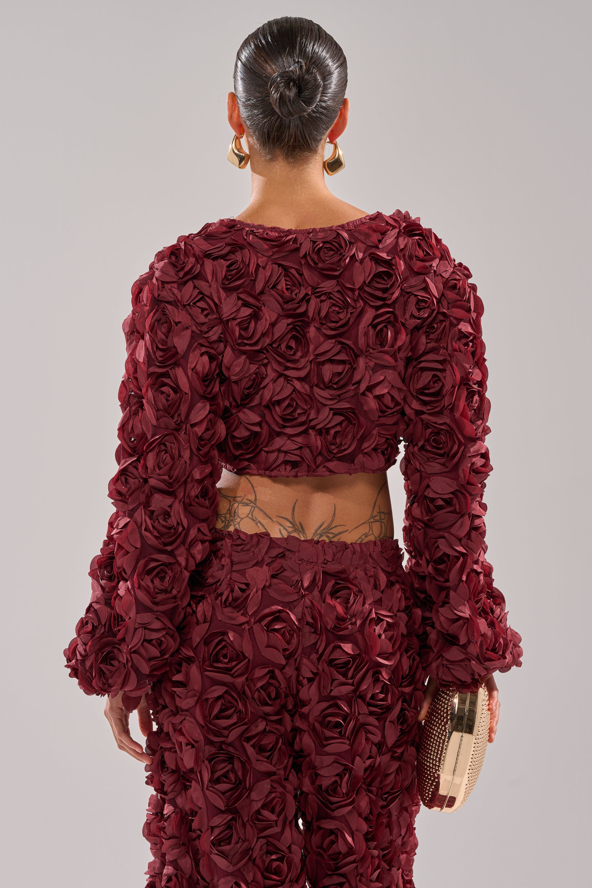 A person with slicked-back hair and gold hoop earrings models the ROSE PETALS FLORAL SHRUG, a dark red cropped jacket with 3D fabric roses. Holding a clutch, they reveal a lower back tattoo. Photo is taken from behind. 3