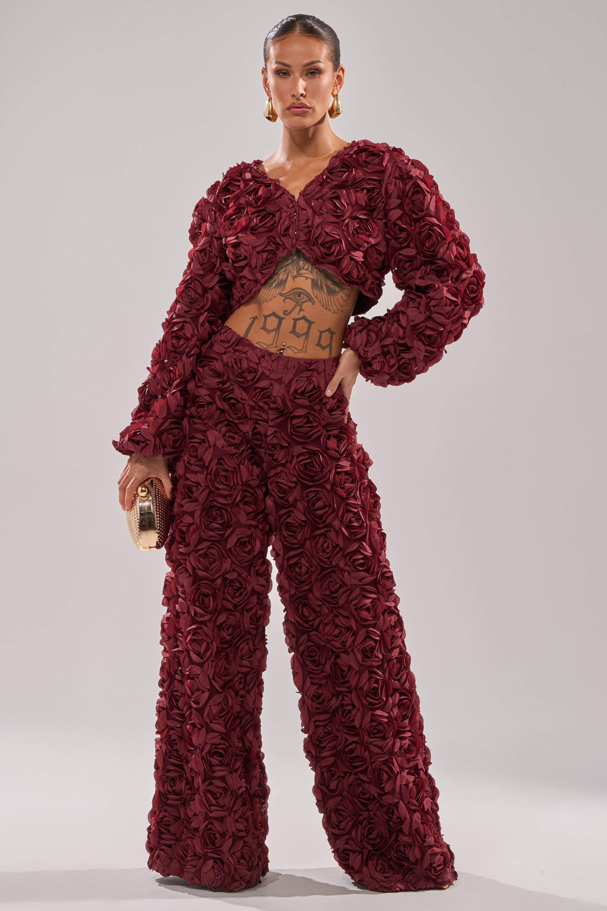 A woman models the ROSE PETALS FLORAL SHRUG, featuring 3D fabric roses in burgundy. She wears it with a matching outfit, slicked-back hair, gold hoop earrings, a tattooed stomach, and holds a gold clutch against a light gray background. 4