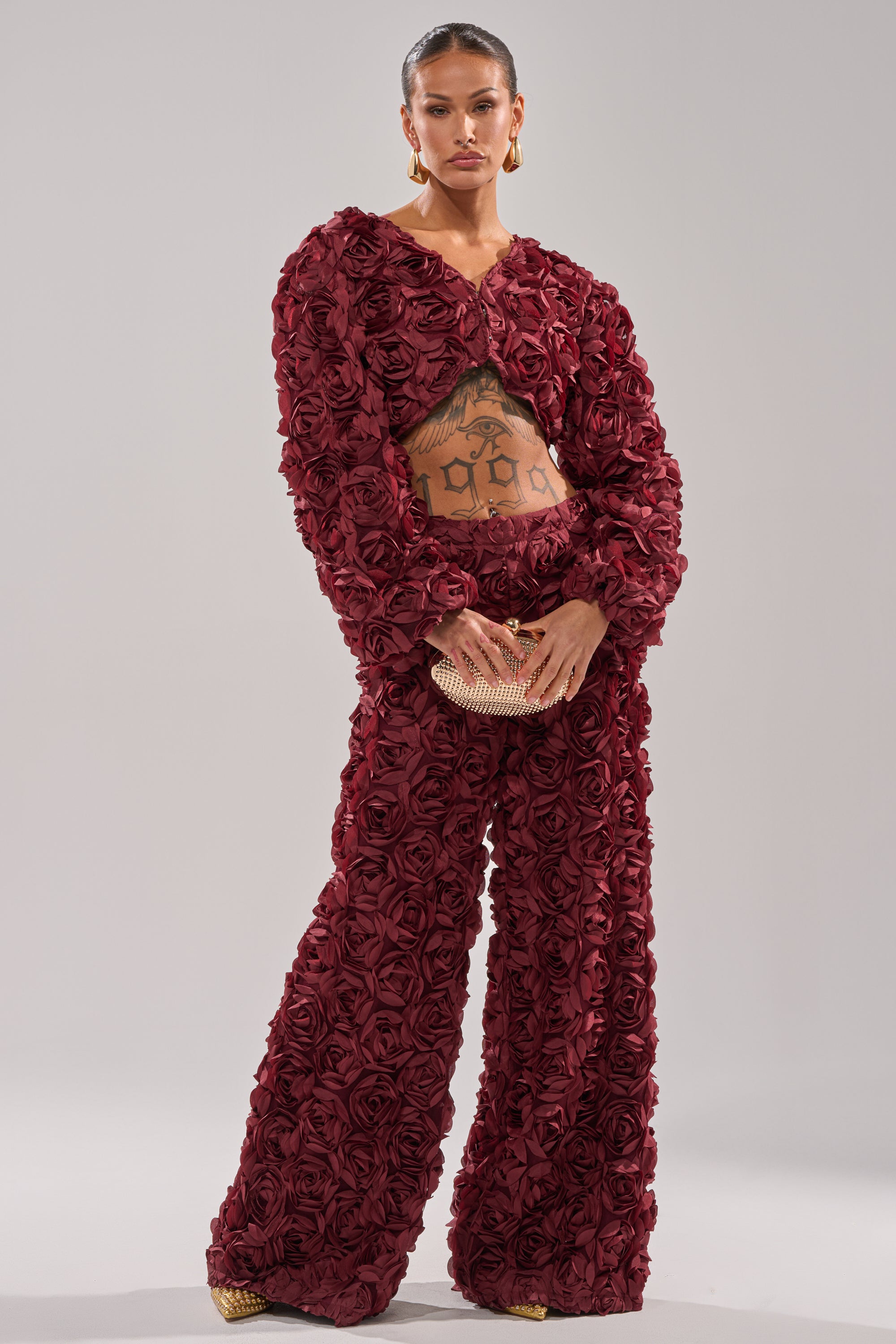 A woman wears the ROSE PETALS FLORAL SHRUG, a burgundy outfit with 3D fabric roses, wide-leg pants, and a cropped jacket. She holds a gold clutch and reveals midriff tattoos while posing against a plain background. 5