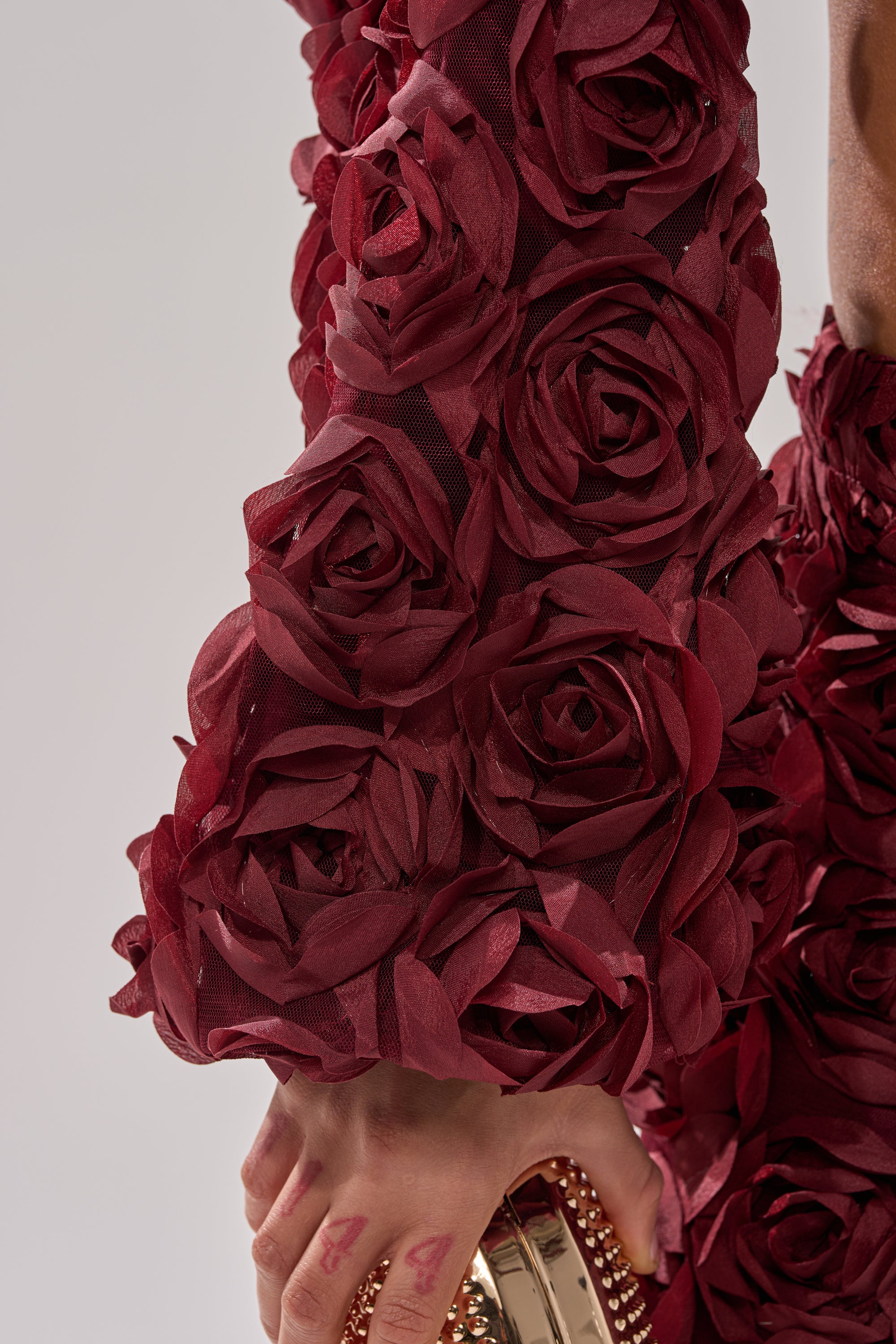 A close-up shows an arm in the ROSE PETALS FLORAL SHRUG with 3D roses, holding a gold studded clutch. Red markings accent the fingers as the deep red textured sleeve stands out against a light gray background. 6