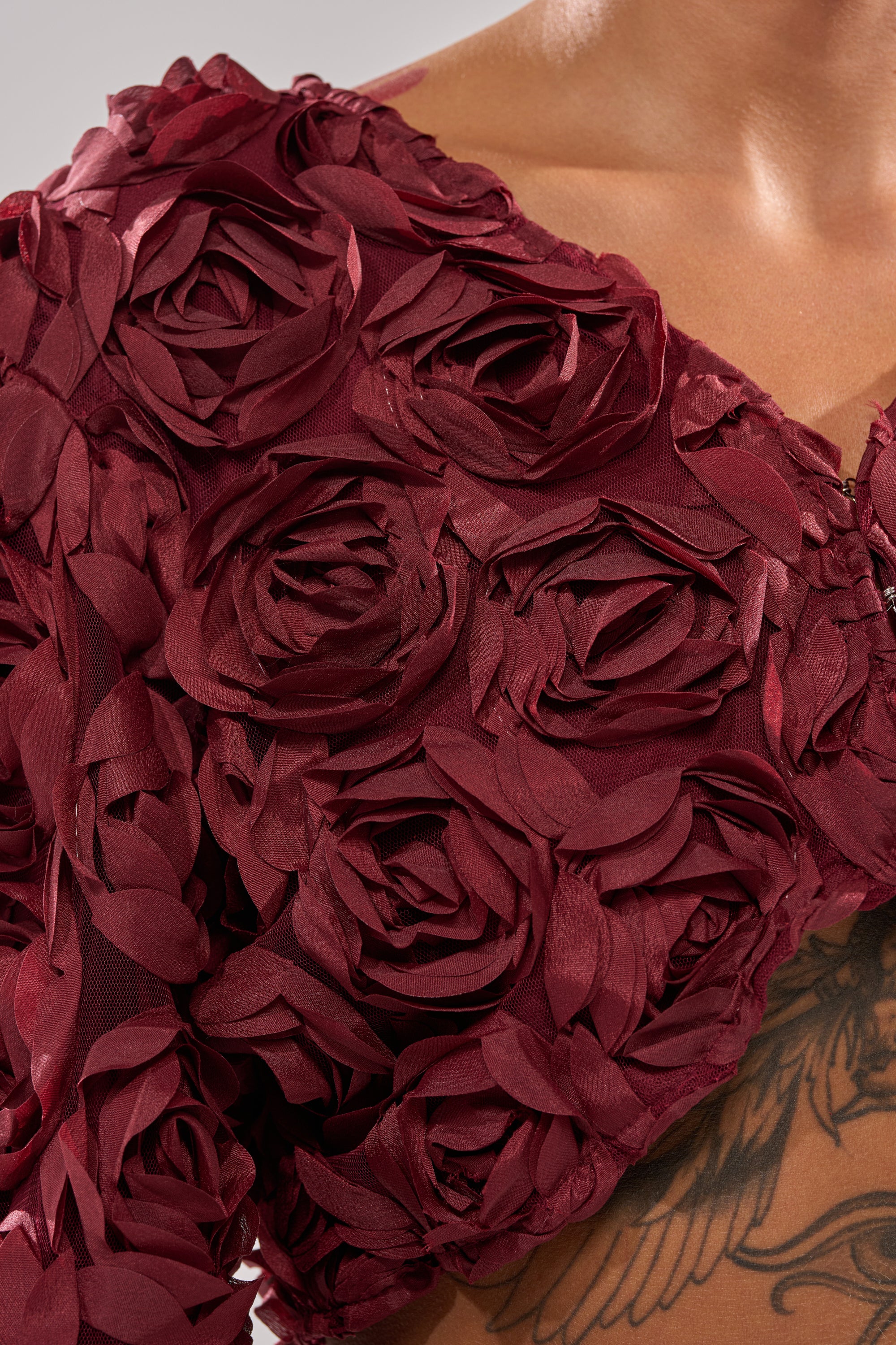 Close-up of a person's upper torso in the ROSE PETALS FLORAL SHRUG, a burgundy top decorated with 3D fabric roses. A tattoo peeks beneath the shrug, while the neutral background highlights its textured floral design. 7