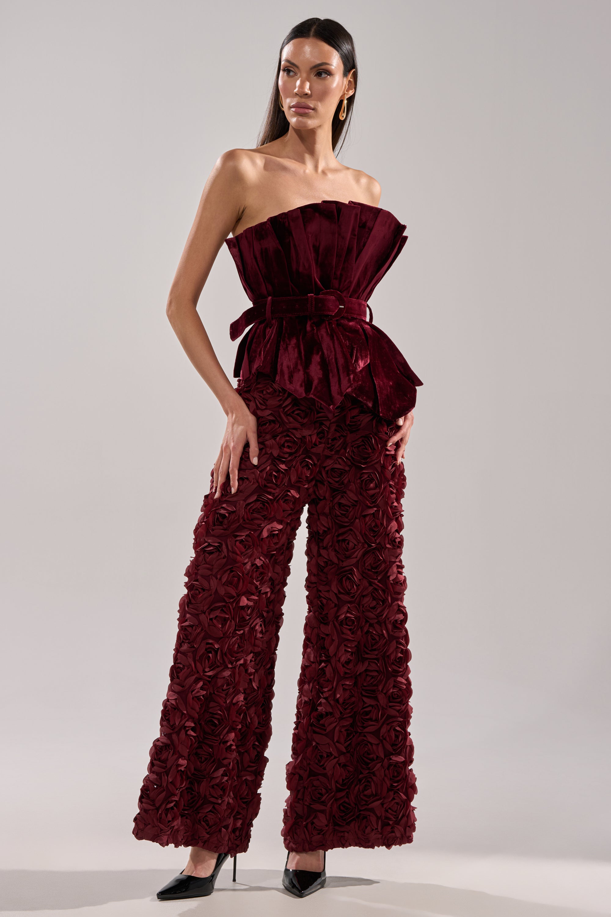 A woman wears the ROSE PEDALS WIDE LEG TROUSER, a high-rise burgundy velvet pant with 3D rose appliques and a matching strapless pleated top, styled with black pointed-toe heels against a plain light background.