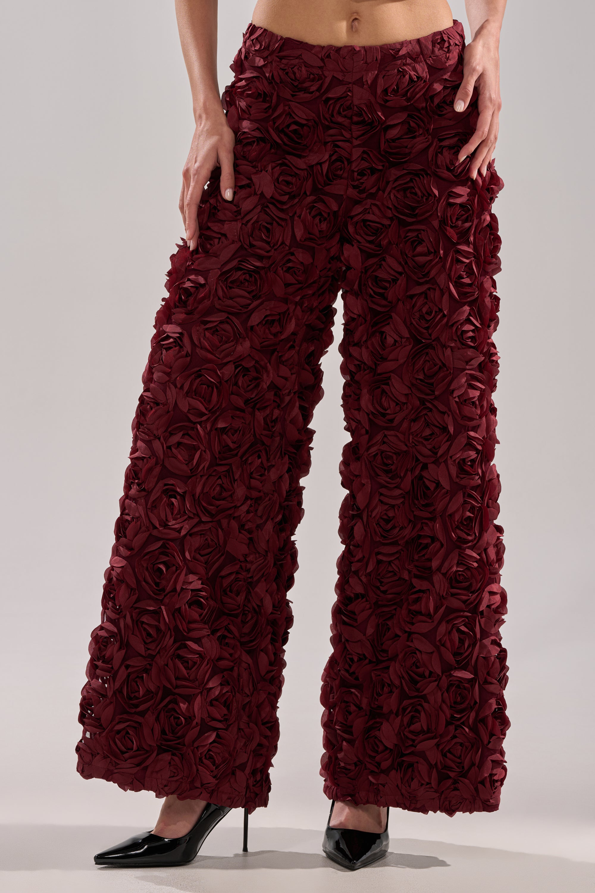 A person wears the ROSE PEDALS WIDE LEG TROUSER in burgundy, featuring textured rose appliques, paired with black pointed high heels. Only the lower torso and legs are shown.