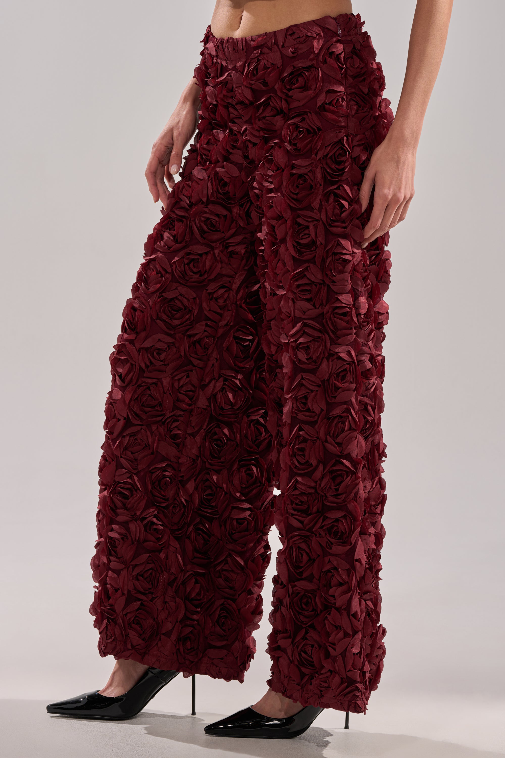 A person wearing the ROSE PEDALS WIDE LEG TROUSER, featuring high-rise, wide-leg design with textured red rose appliqués, paired with black pointed-toe heels.