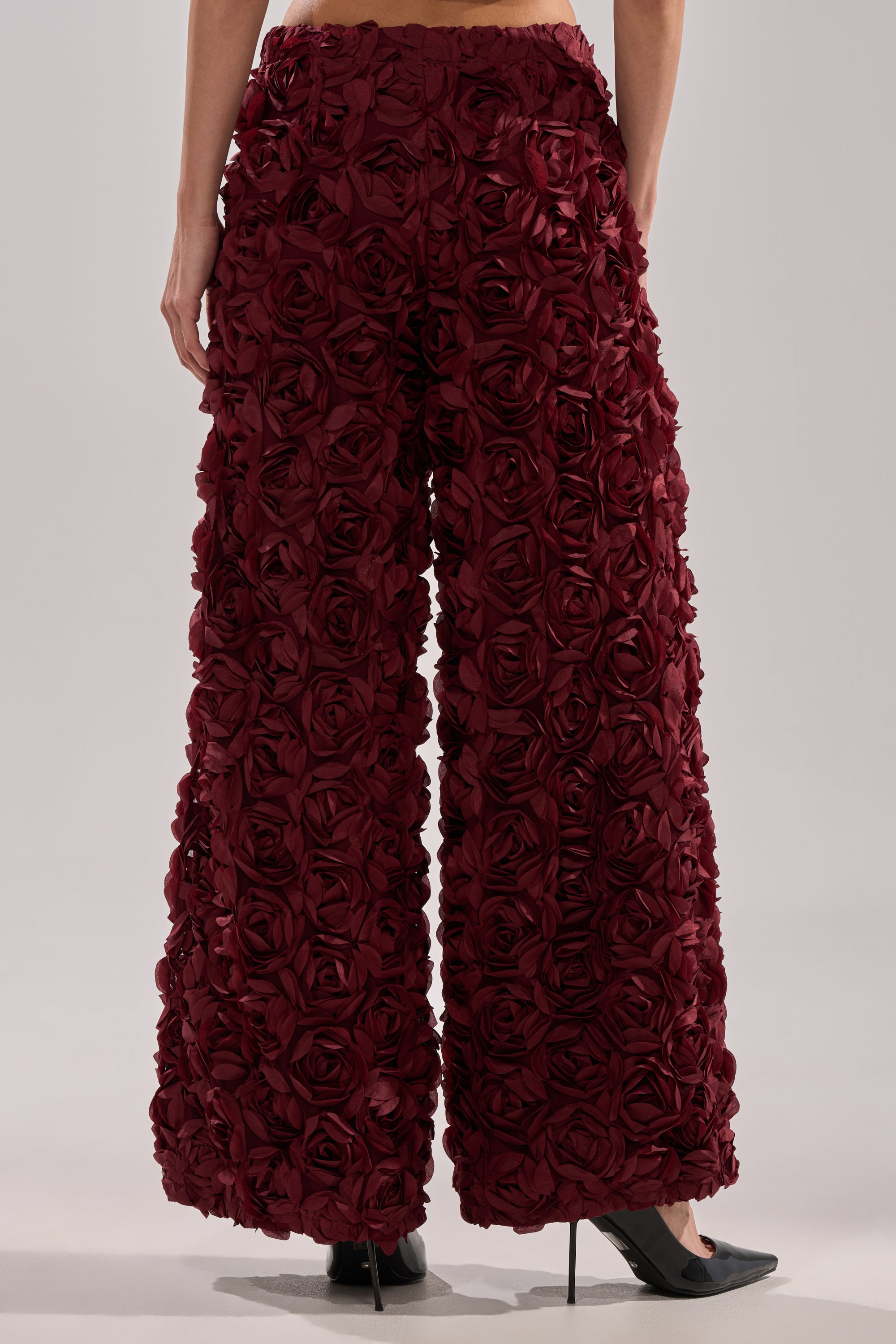 A person stands facing away, wearing the ROSE PEDALS WIDE LEG TROUSER featuring three-dimensional rose appliques, paired with black pointed-toe heels.
