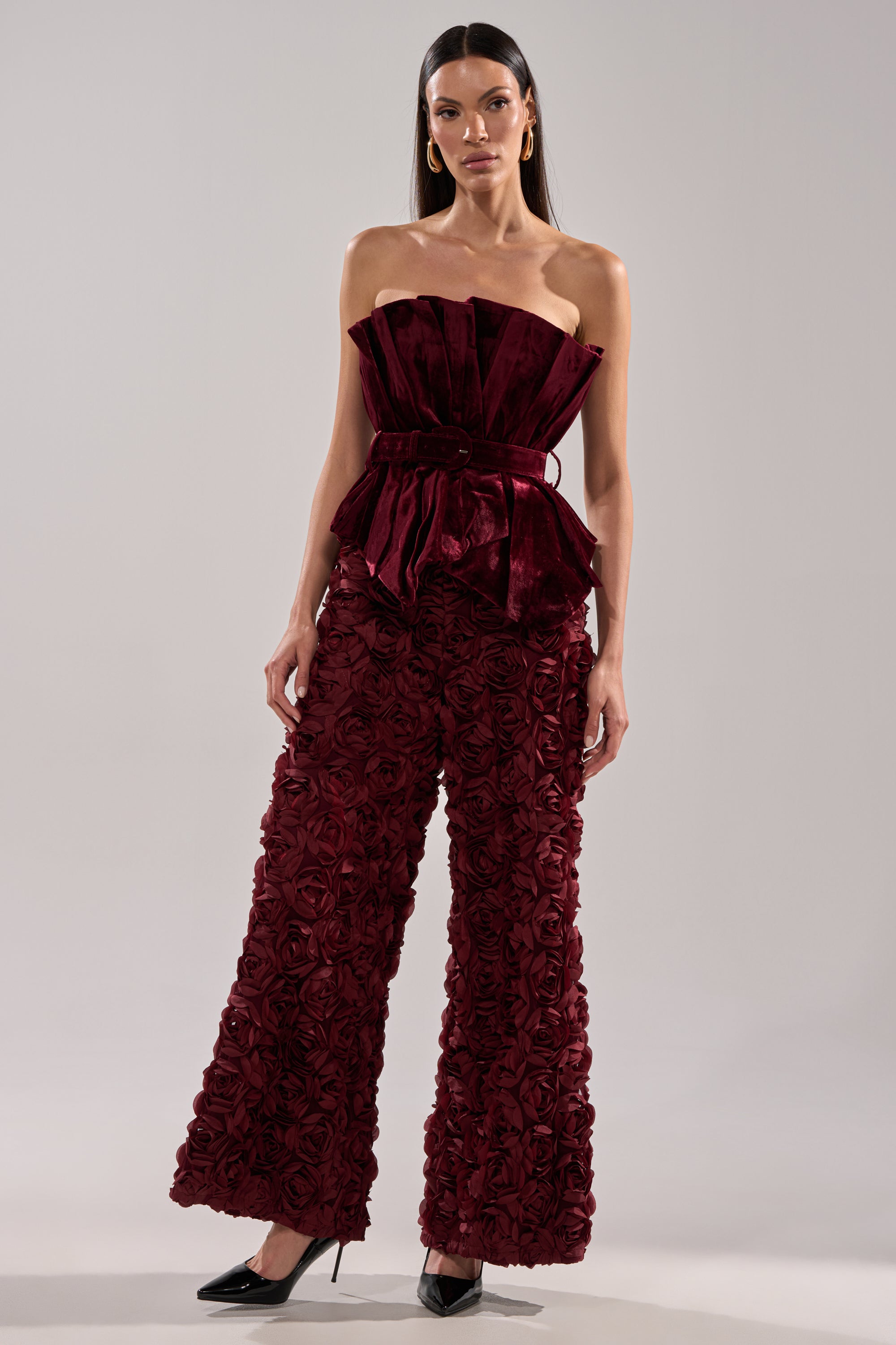 A woman wears the ROSE PEDALS WIDE LEG TROUSER, styled with a strapless deep red velvet top featuring a structured ruffled bodice. She finishes the look with black pointed heels.
