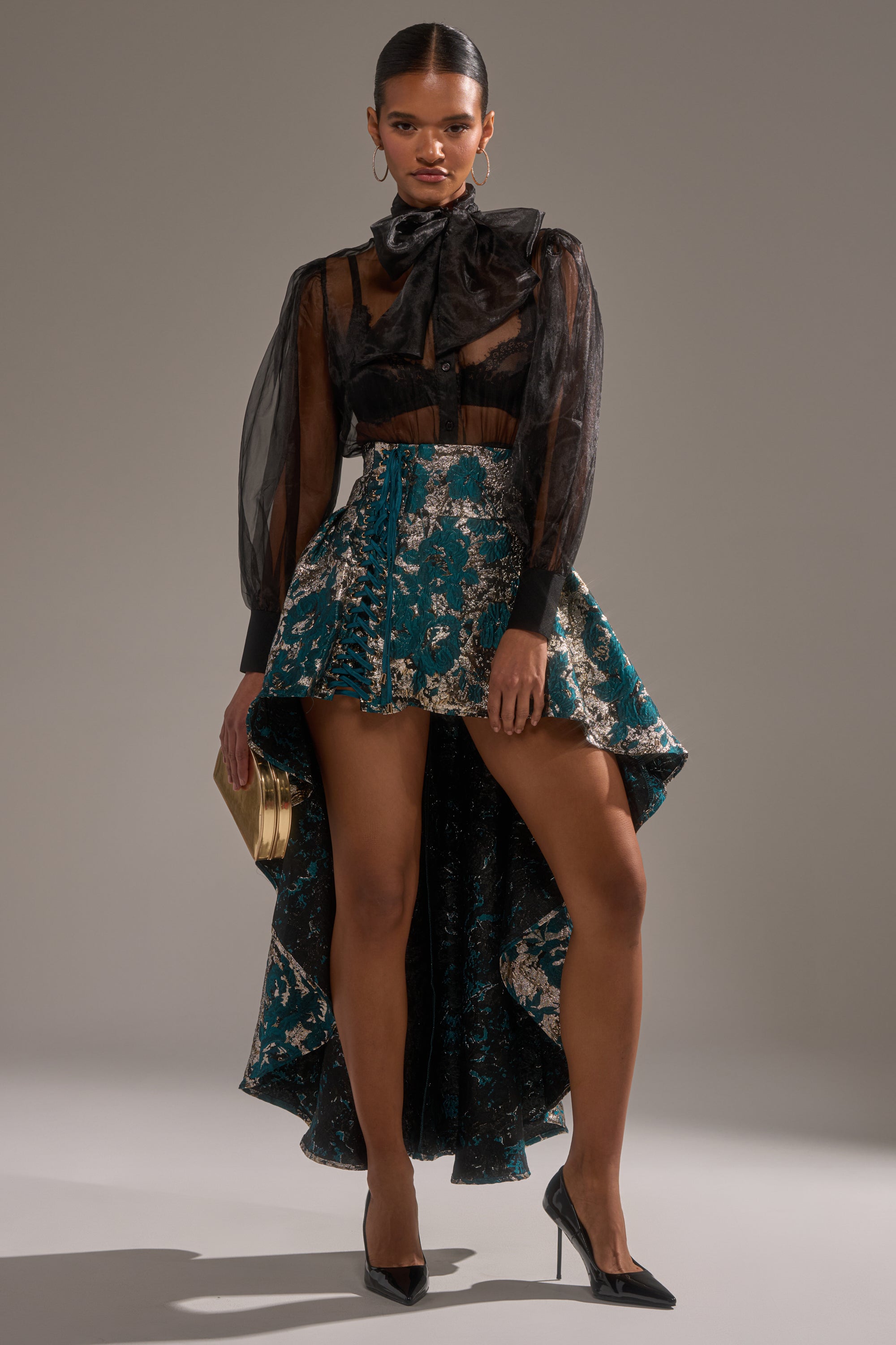 A woman stands confidently against a neutral background, wearing the GLORIOUS DAYS BROCADE HIGH LOW SKIRT in teal and gold with a sheer black blouse, large bow, black pointed heels, and a gold clutch.