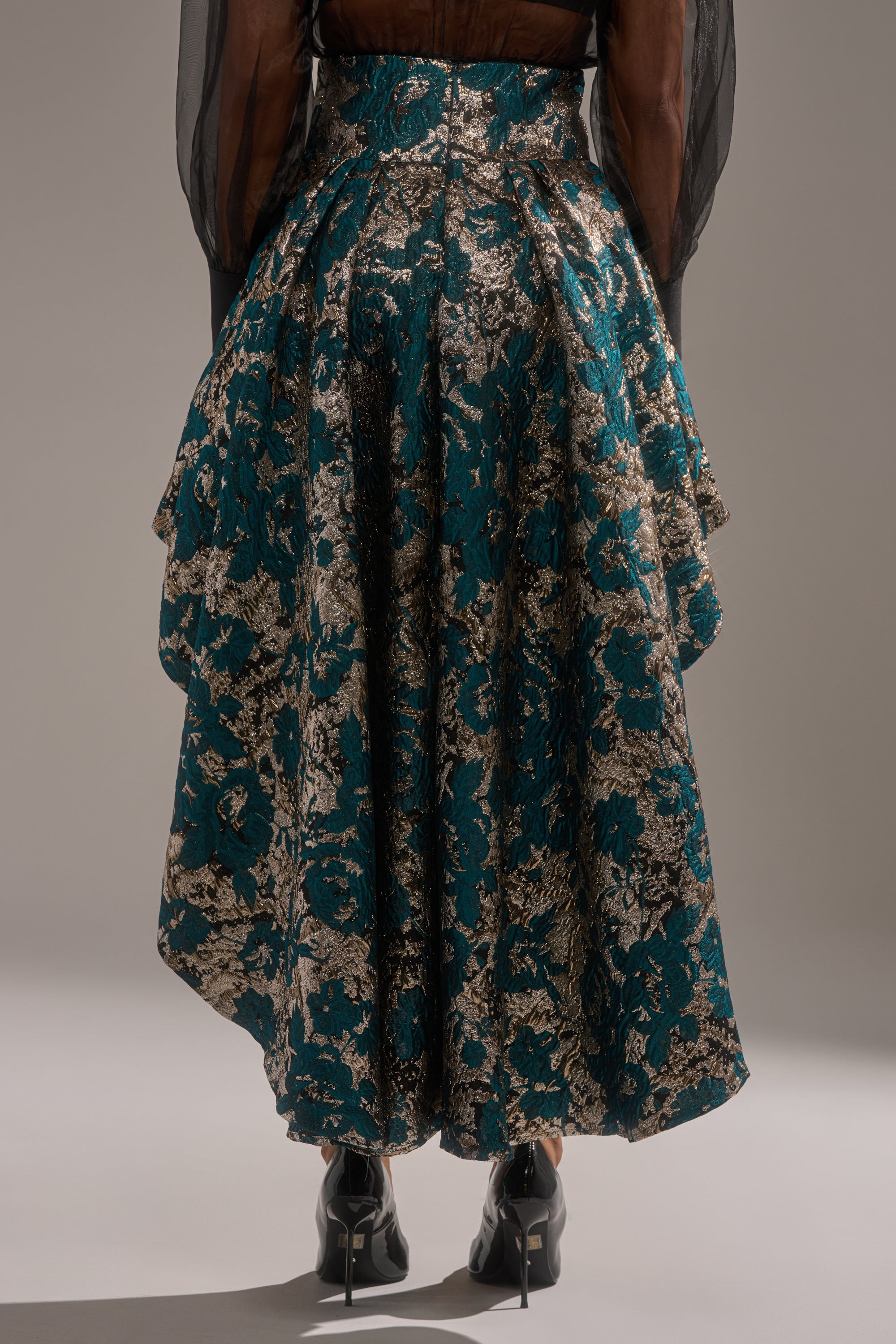 A person stands facing away, wearing the GLORIOUS DAYS BROCADE HIGH LOW SKIRT with metallic teal and gold brocade, ruffled layers, black heeled shoes, and a sheer black long-sleeve top.