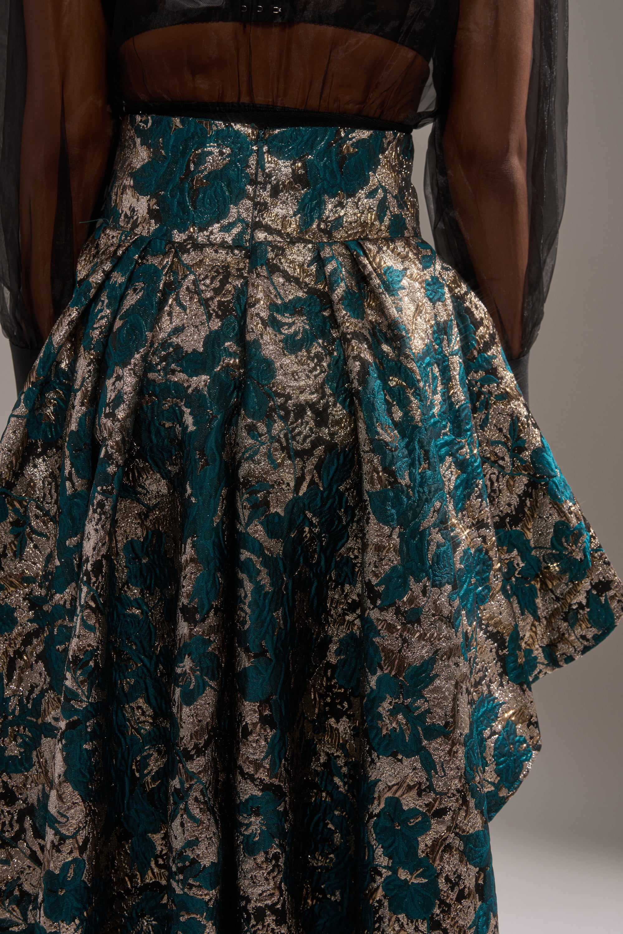 A close-up of a person wearing a sheer black blouse with long sleeves and the GLORIOUS DAYS BROCADE HIGH LOW SKIRT, shown from the back against a plain background.