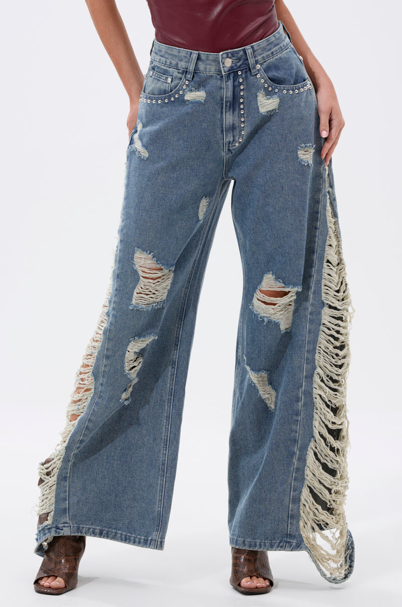 Someone wearing the OVER IT DISTRESSED RELAXED DENIM—wide-leg jeans with large rips and fringe on the side—styled with a sleeveless maroon top and open-toe heels.
