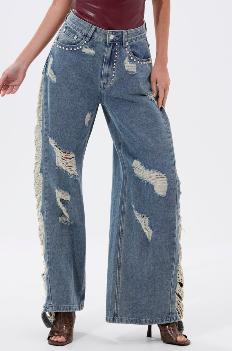 Someone wearing the OVER IT DISTRESSED RELAXED DENIM—high-rise, wide-leg jeans with rips and distressed patches—paired with a maroon top and open-toe heels.