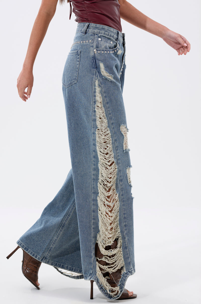 A woman wears the OVER IT DISTRESSED RELAXED DENIM, high-rise wide-leg jeans with dramatic rips and frayed edges, styled with a sleeveless burgundy top and brown heeled sandals.