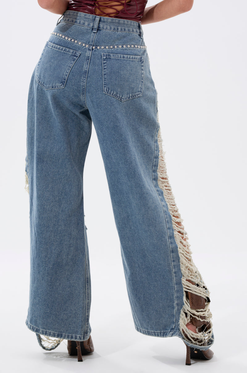 A person is seen from the back wearing OVER IT DISTRESSED RELAXED DENIM, featuring high-rise, wide legs, and heavy distressing, paired with heeled shoes against a plain background.