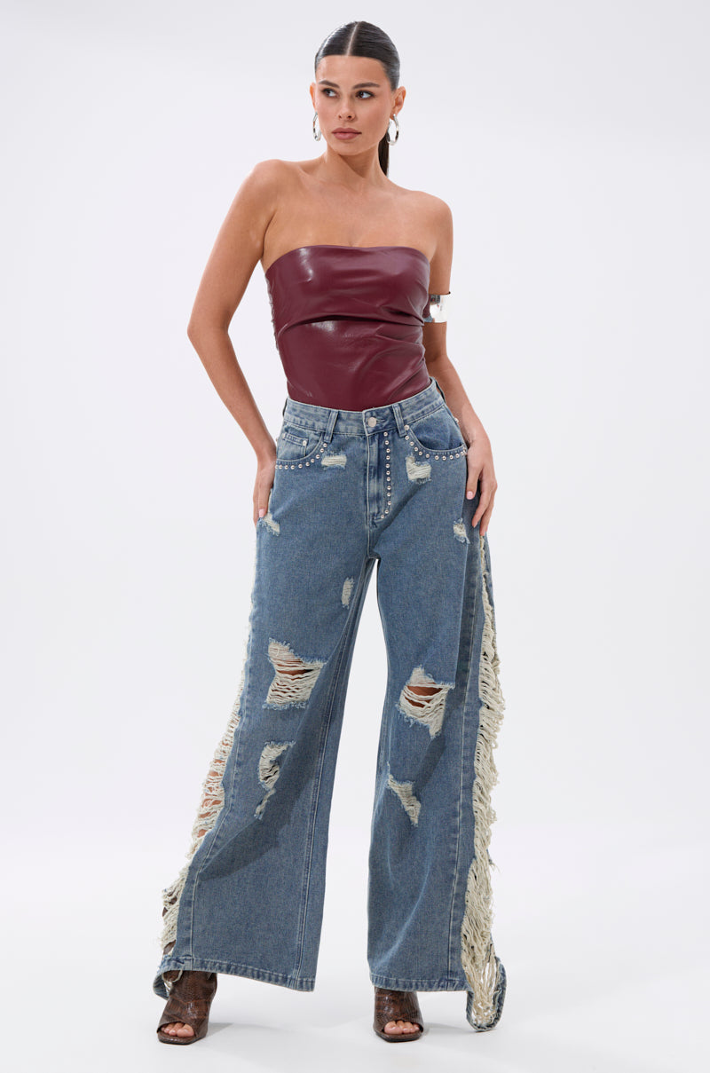 A woman poses in the OVER IT DISTRESSED RELAXED DENIM, featuring wide legs with heavy rips and fringe on the sides, paired with a maroon strapless leather top, heeled sandals, and a sleek ponytail.