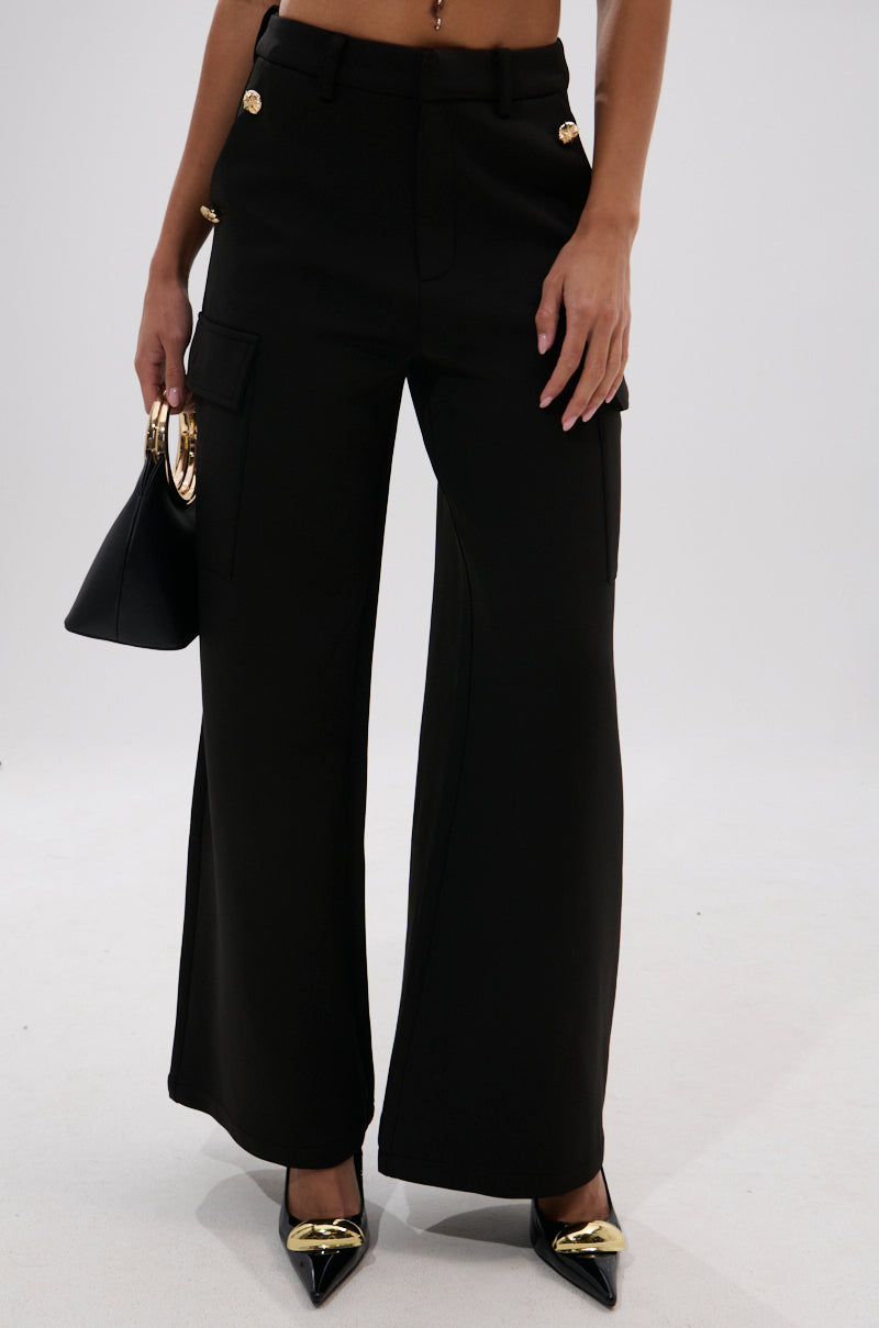 Wearing the GOTTA LET IT GO TROUSER, high-rise and black with gold buttons, paired with pointed black heels featuring gold tips, and carrying a small black handbag.