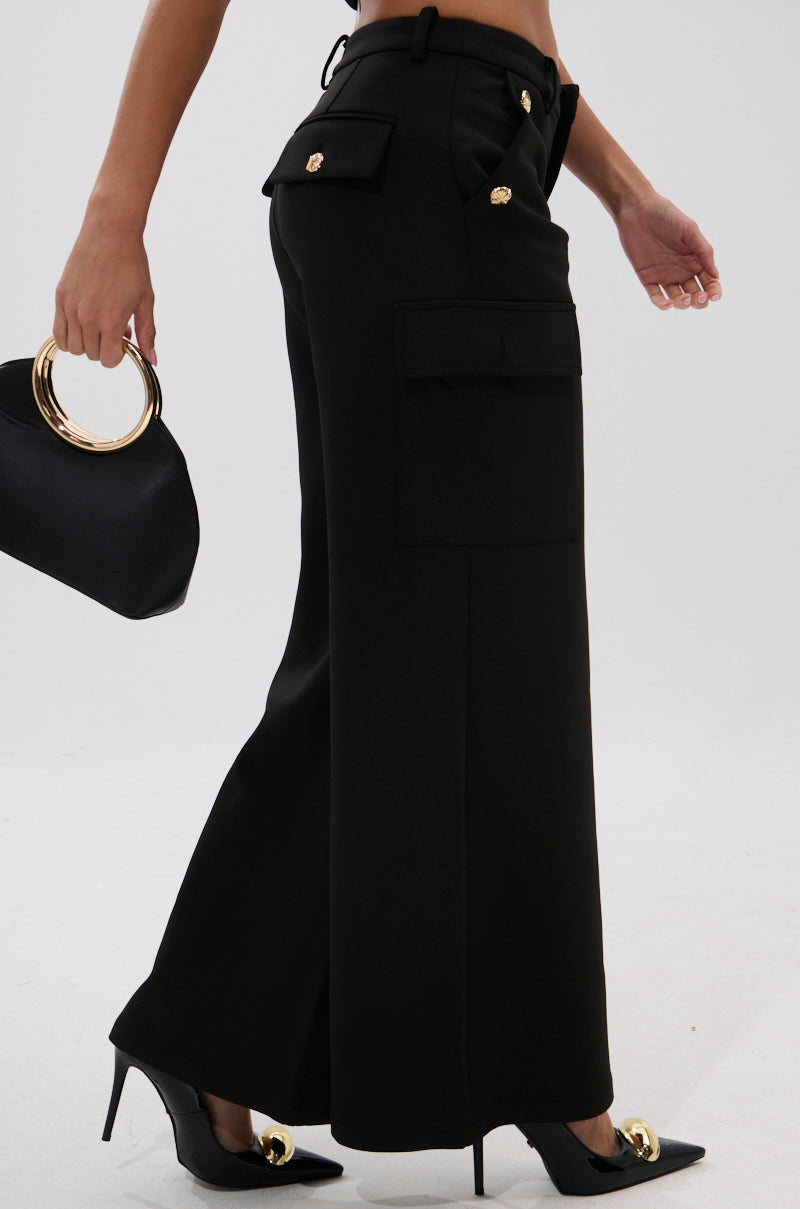 A person wears GOTTA LET IT GO TROUSER—high-rise black wide-leg trousers with gold buttons—paired with black pointed-toe heels with gold accents and a black handbag with a gold circular handle, set against a plain white background.