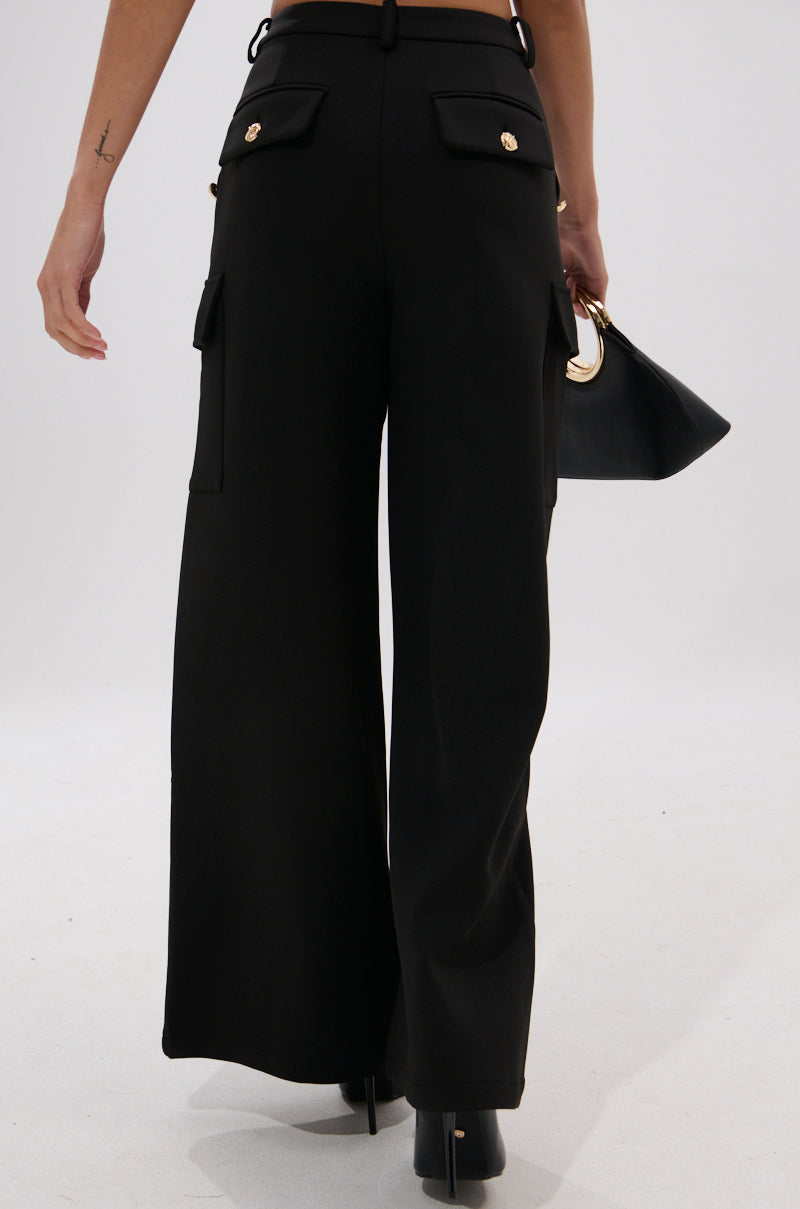 A person is shown from the back wearing the GOTTA LET IT GO TROUSER, which features high-rise wide legs, cargo flap pockets, and gold buttons. They hold a black handbag with a gold handle against a plain light background.