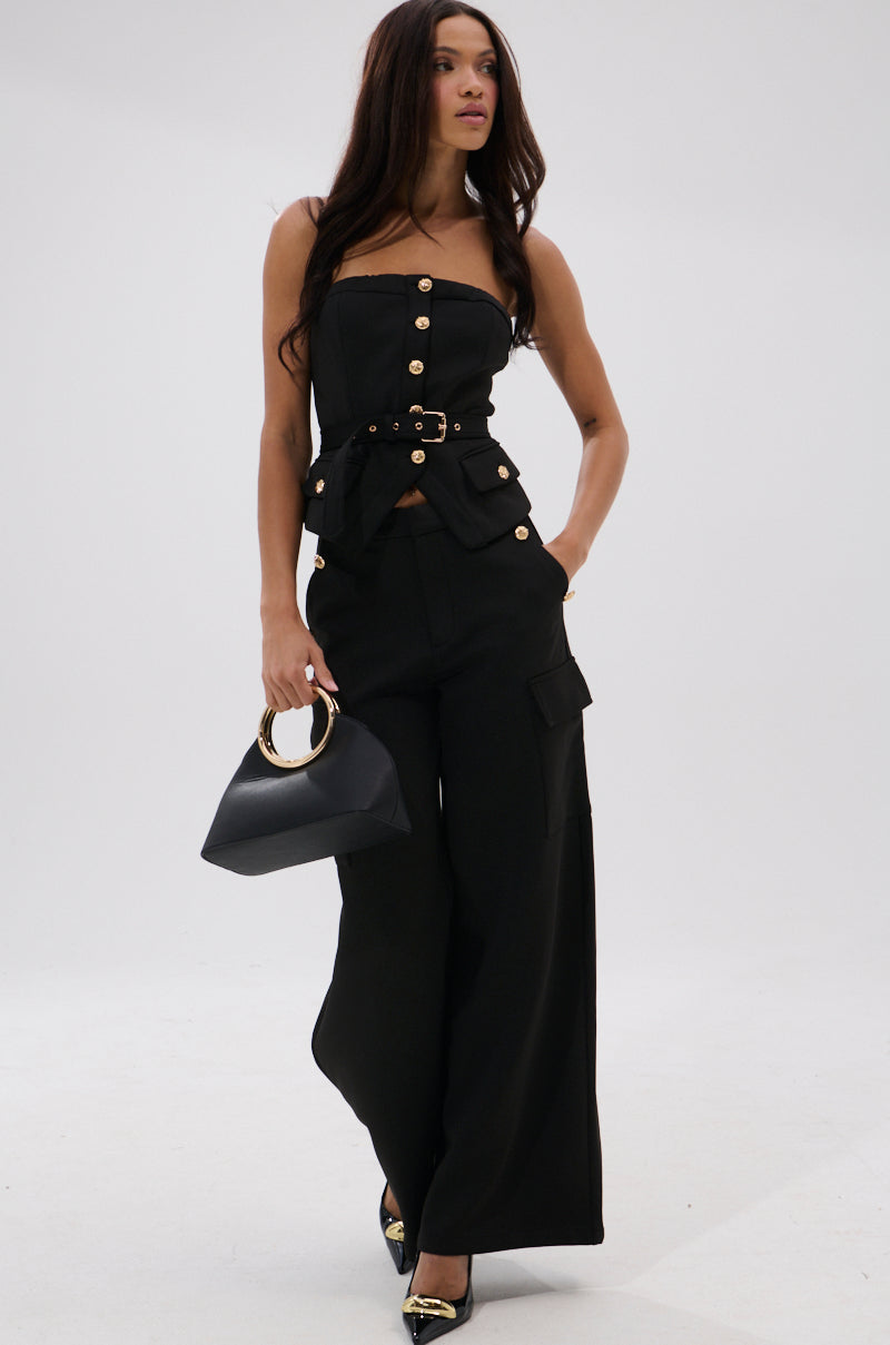 A woman in the GOTTA LET IT GO TROUSER, a black strapless top with gold buttons, black heels, and a black handbag with a round gold handle walks forward confidently.