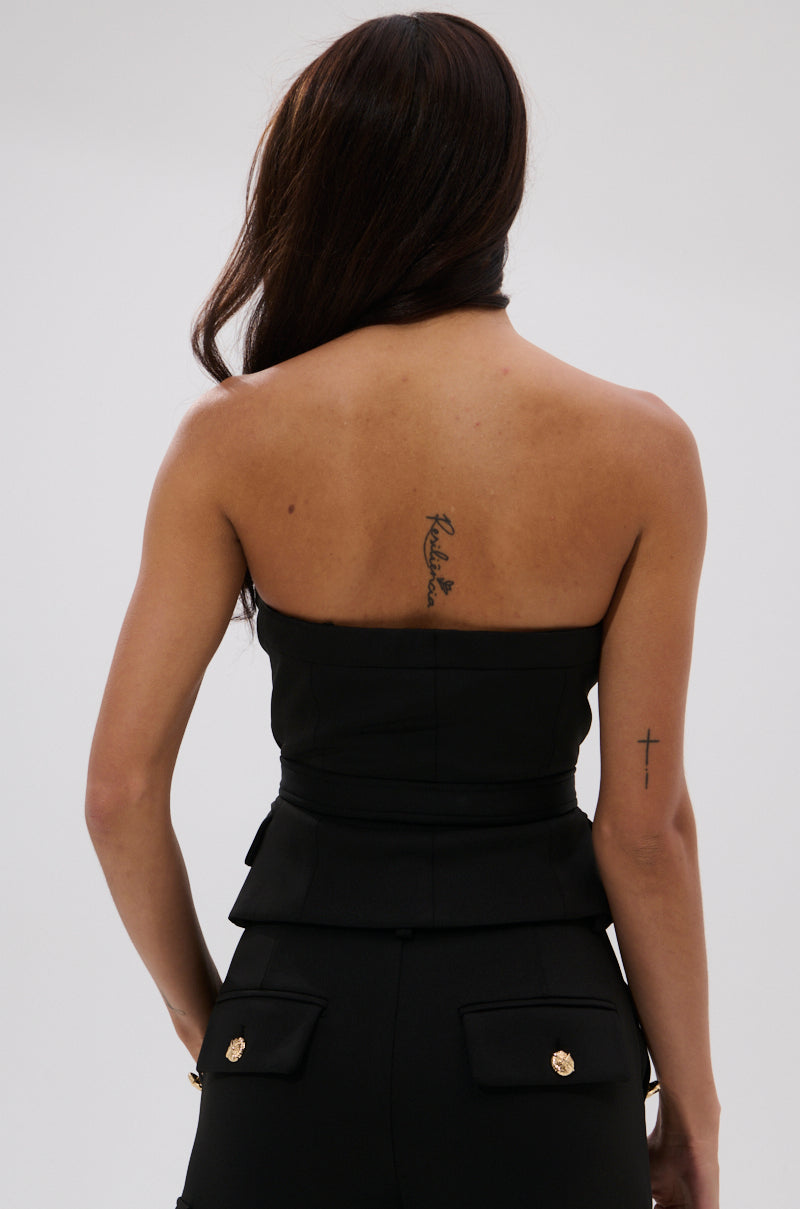 A woman with long brown hair and tattoos on her upper back and left arm faces away, wearing the GOTTA LET IT GO BUCKLE CORSET and matching black pants with gold buttons. She stands against a plain, light background.
