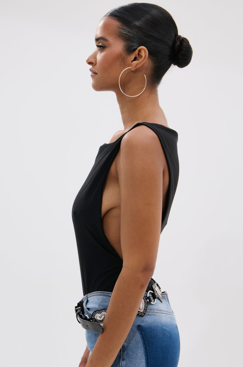 A woman with dark hair in a sleek bun, large hoop earrings, and blue jeans with a decorative belt stands in profile against a plain background, modeling the SEXY OPEN SIDE BODYSUIT featuring deep armholes and a curve-skimming fit.
