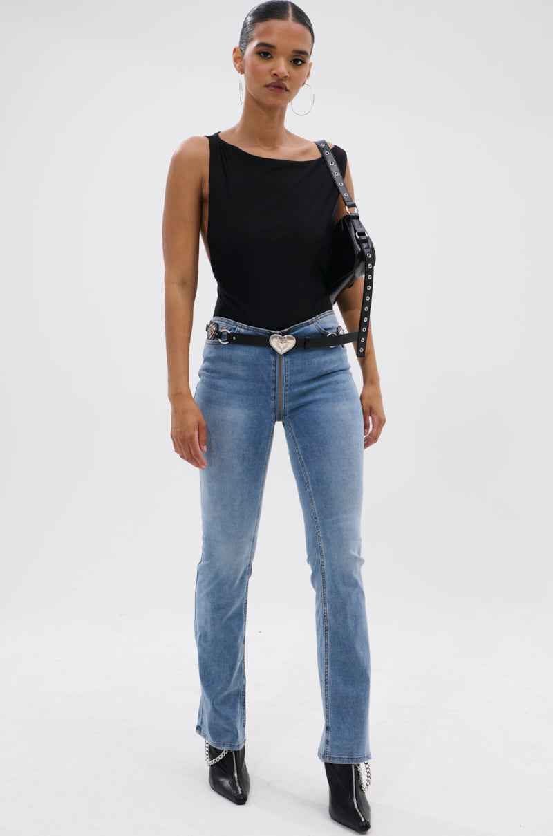 A woman stands against a white background wearing the SEXY OPEN SIDE BODYSUIT, paired with high-waisted light blue jeans featuring a heart-shaped belt buckle, black boots, hoop earrings, and a black shoulder bag.