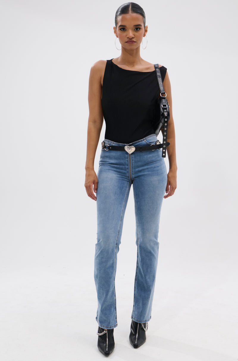A woman poses against a white background in the SEXY OPEN SIDE BODYSUIT, paired with light blue jeans featuring a heart-shaped belt buckle, black pointed-toe boots, and a black strap over her shoulder.