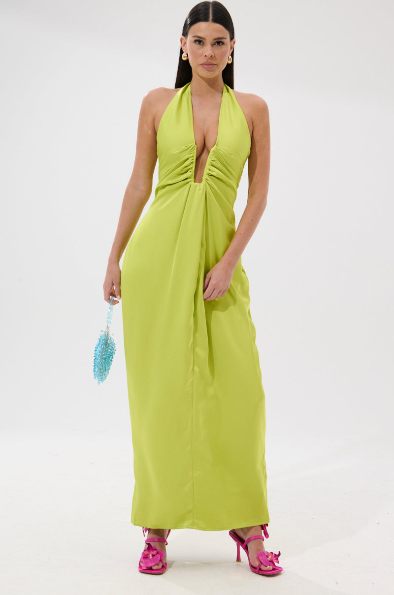 A woman poses against a white backdrop in the IN YOUR DREAMS SATIN MIDI DRESS, featuring a lime green halter style with a deep V neckline. She accessorizes with a blue beaded purse and hot pink high-heeled sandals.