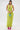 A woman with long, straight dark hair and a neutral expression stands against a white background wearing the IN YOUR DREAMS SATIN MIDI DRESS in lime green, featuring a sleeveless halter neckline. She pairs it with pink heels and a blue beaded handbag.