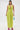 A woman poses against a white backdrop in the IN YOUR DREAMS SATIN MIDI DRESS, a sleeveless lime green halter dress with a deep V neckline. She accessorizes with a small blue beaded bag and bright pink heeled sandals.
