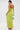 Woman facing away in the IN YOUR DREAMS SATIN MIDI DRESS in bright lime green, holding a small blue beaded handbag and wearing pink high-heeled sandals. Background is plain white.