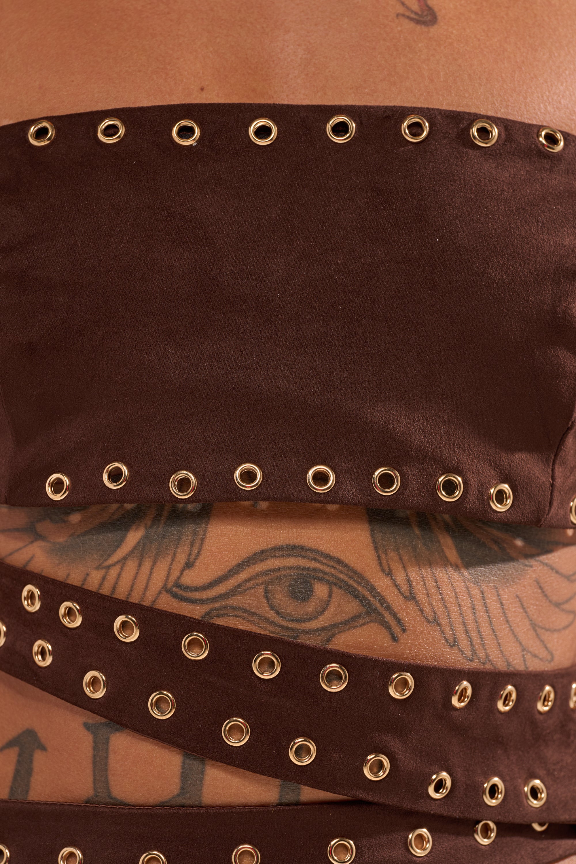 Close-up of a torso in the PARTYS HERE SUEDE CUTOUT MINI DRESS with gold eyelets, partially revealing a winged eye tattoo underneath. 9