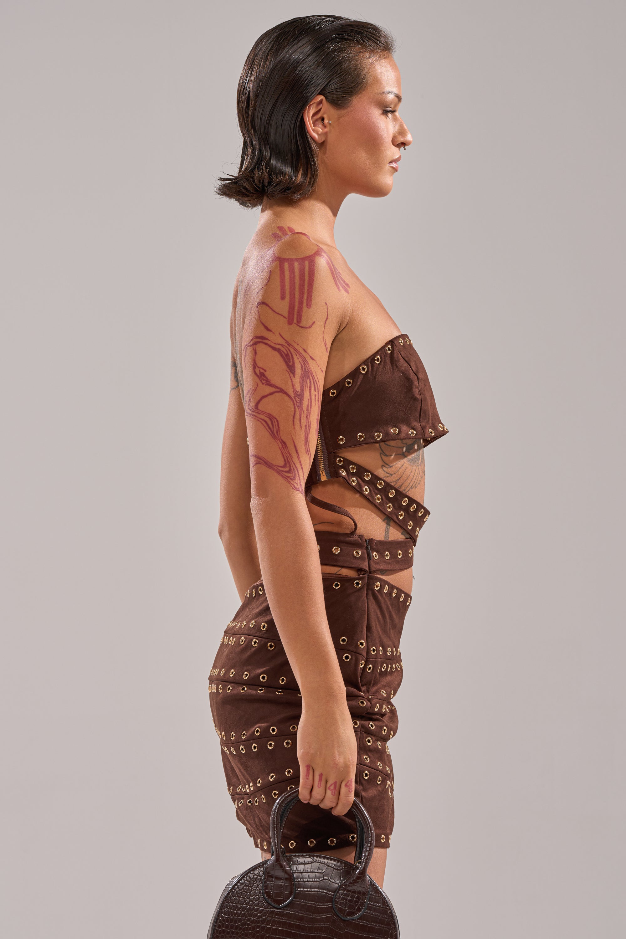 A person with short dark hair and abstract tattoos on their right arm and shoulder stands in profile, wearing the PARTY'S HERE SUEDE CUTOUT MINI DRESS—brown with grommets and side cutouts—and holding a dark-textured handbag. 2