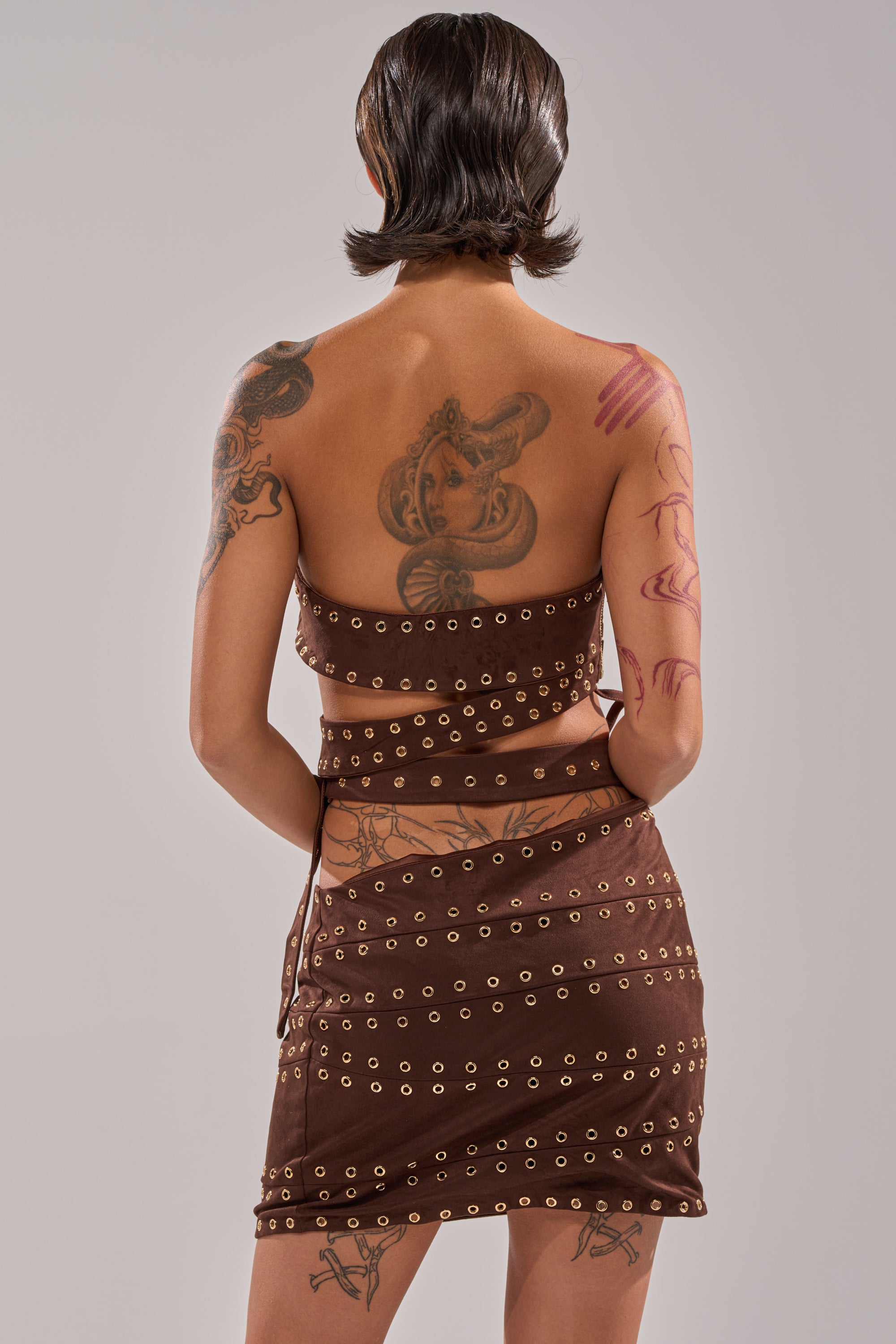 A person with short, slicked-back hair is seen from behind wearing the PARTYS HERE SUEDE CUTOUT MINI DRESS with gold eyelets. Tattoos, including a large snake and abstract designs, cover their arms and upper back. 3