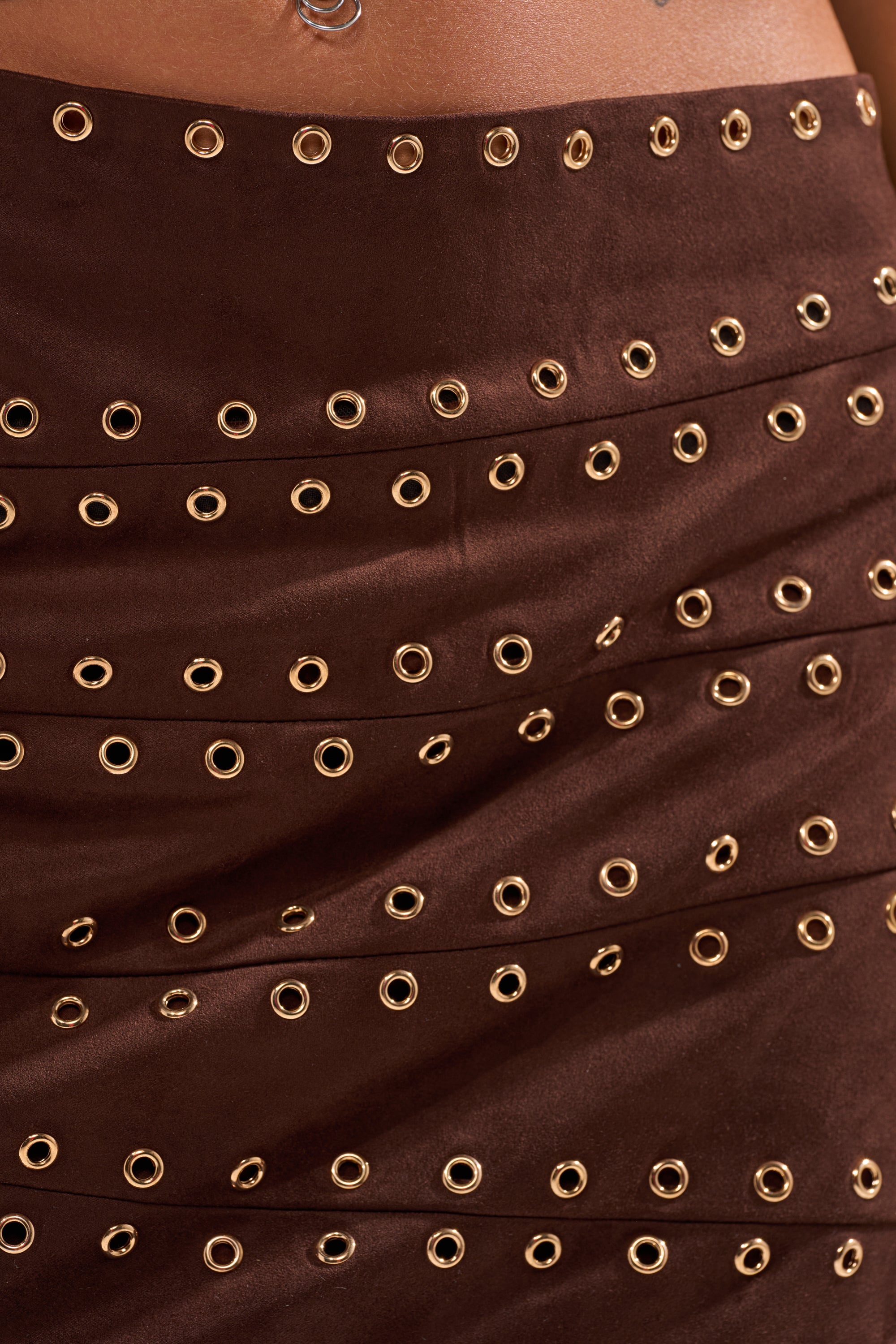 Close-up of the PARTYS HERE SUEDE CUTOUT MINI DRESS, featuring a brown suede skirt detailed with rows of gold eyelets that form a striking textured metallic pattern. 7