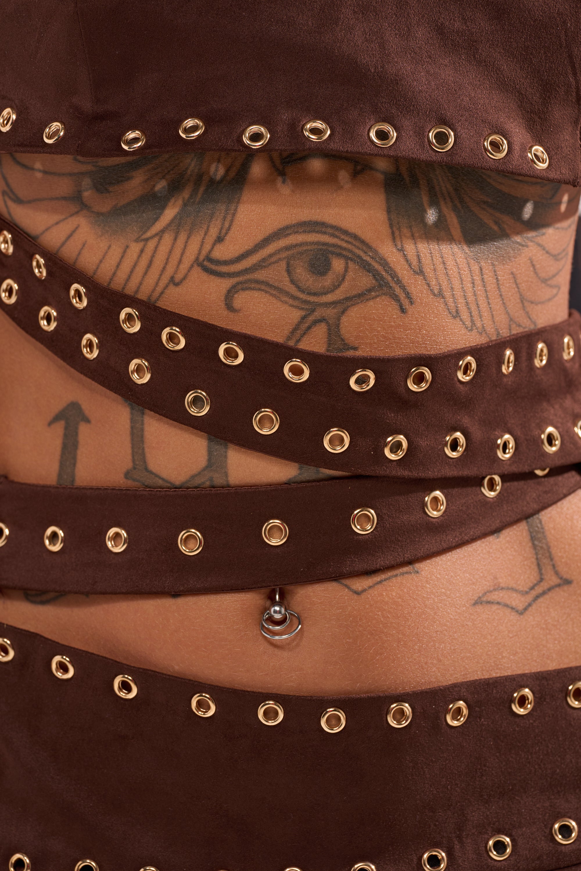 Close-up of a torso with an Eye of Horus tattoo, wearing brown fabric bands with gold eyelets over the PARTYS HERE SUEDE CUTOUT MINI DRESS, accessorized with a navel piercing. 8