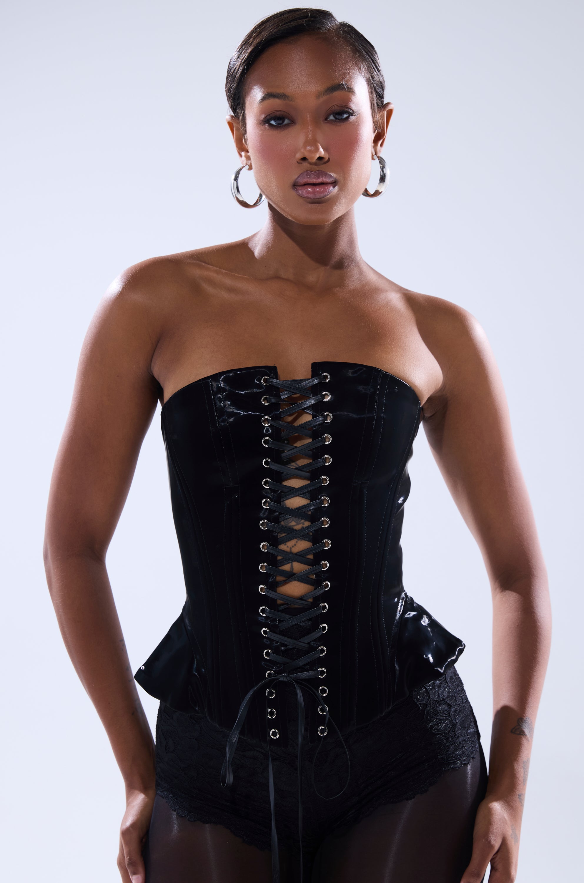 A woman stands confidently in the SMOOTH TRANSITION LACE UP CORSET, styled with lace shorts and large hoop earrings. Her hair is slicked back, and she maintains a neutral expression against a plain background.
