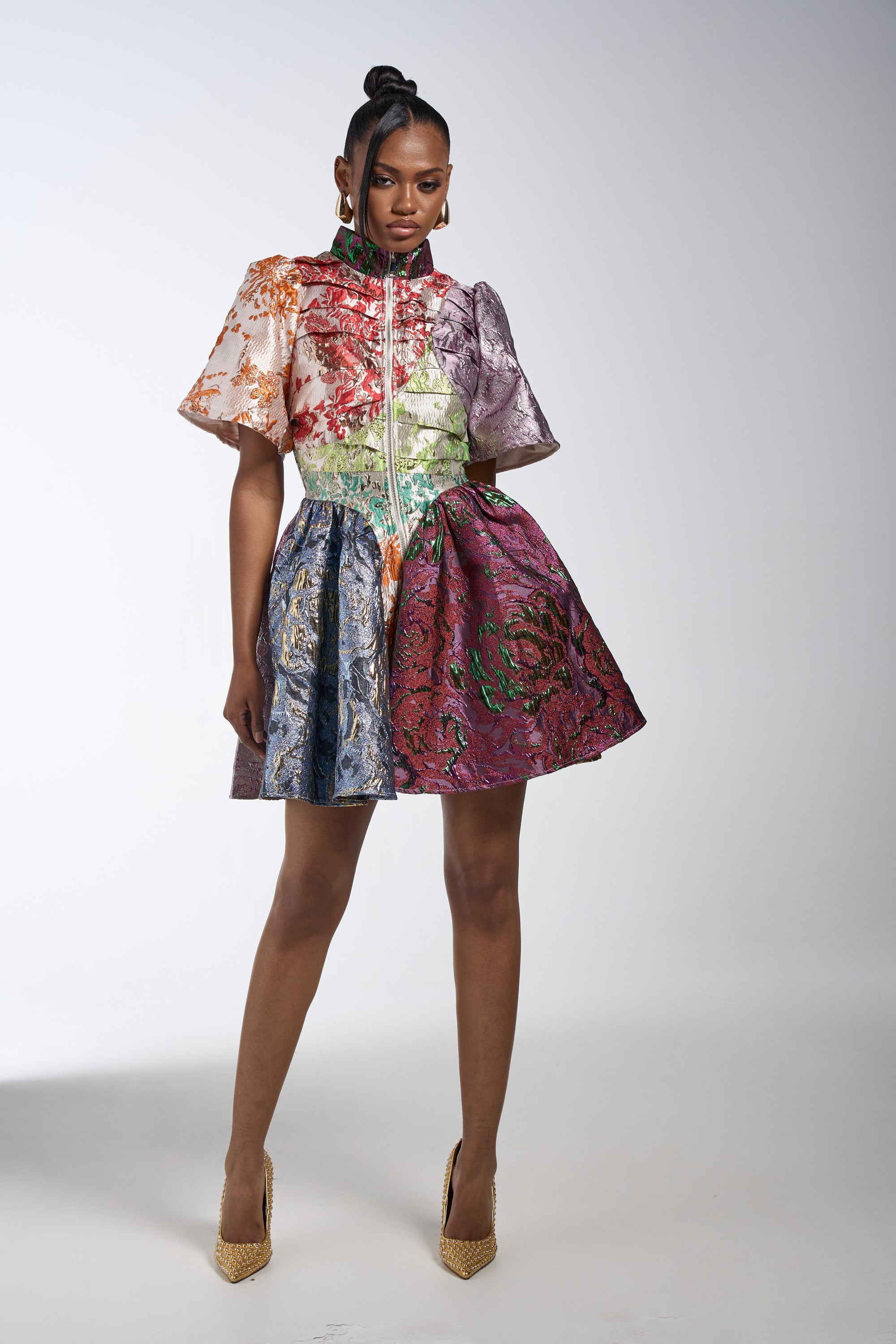 A woman poses confidently against a white background in the STAY WITH ME BROCADE MINI DRESS by AKIRA Label, featuring a fit-and-flare silhouette, short sleeves, and front zipper. She styles it with gold high heels and earrings.