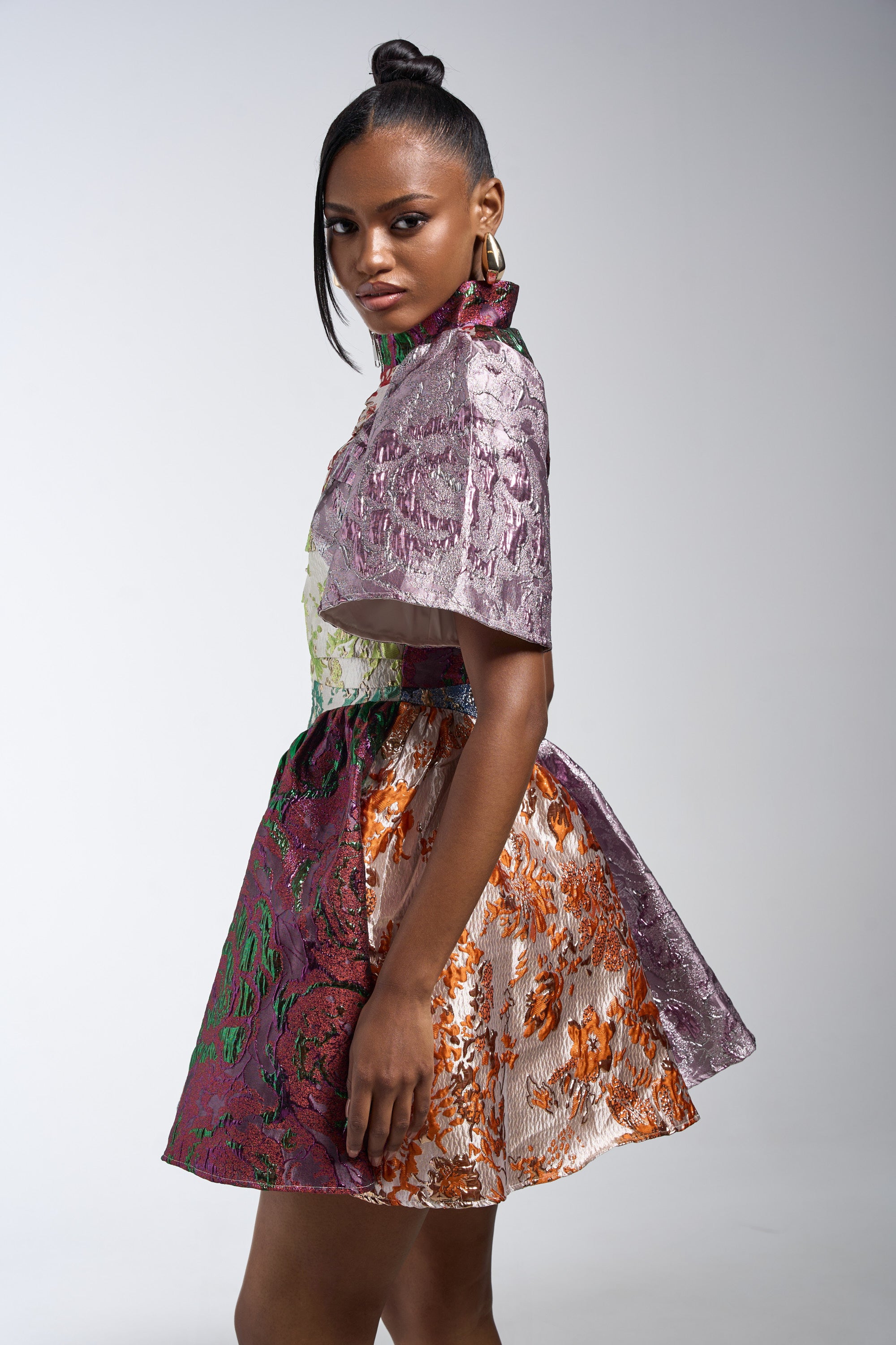 A woman models the STAY WITH ME BROCADE MINI DRESS, a floral jacquard piece with short sleeves and a flared skirt. She pairs it with hoop earrings and a top knot bun, set against a plain light background.