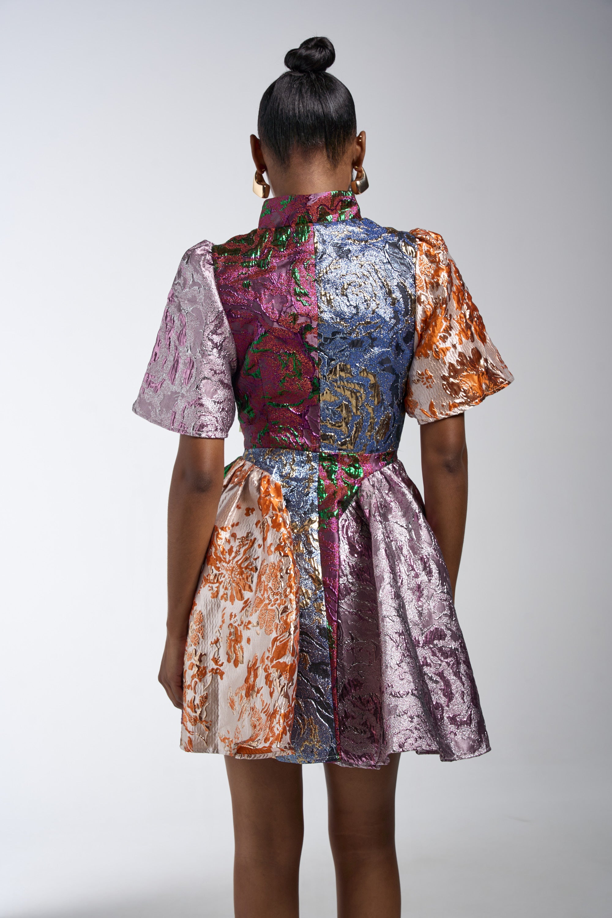 A woman, hair in a neat bun, faces away while wearing the STAY WITH ME BROCADE MINI DRESS—a patchwork mini with metallic purple, silver, orange, and blue floral patterns, and a shiny finish. She stands against a plain background.