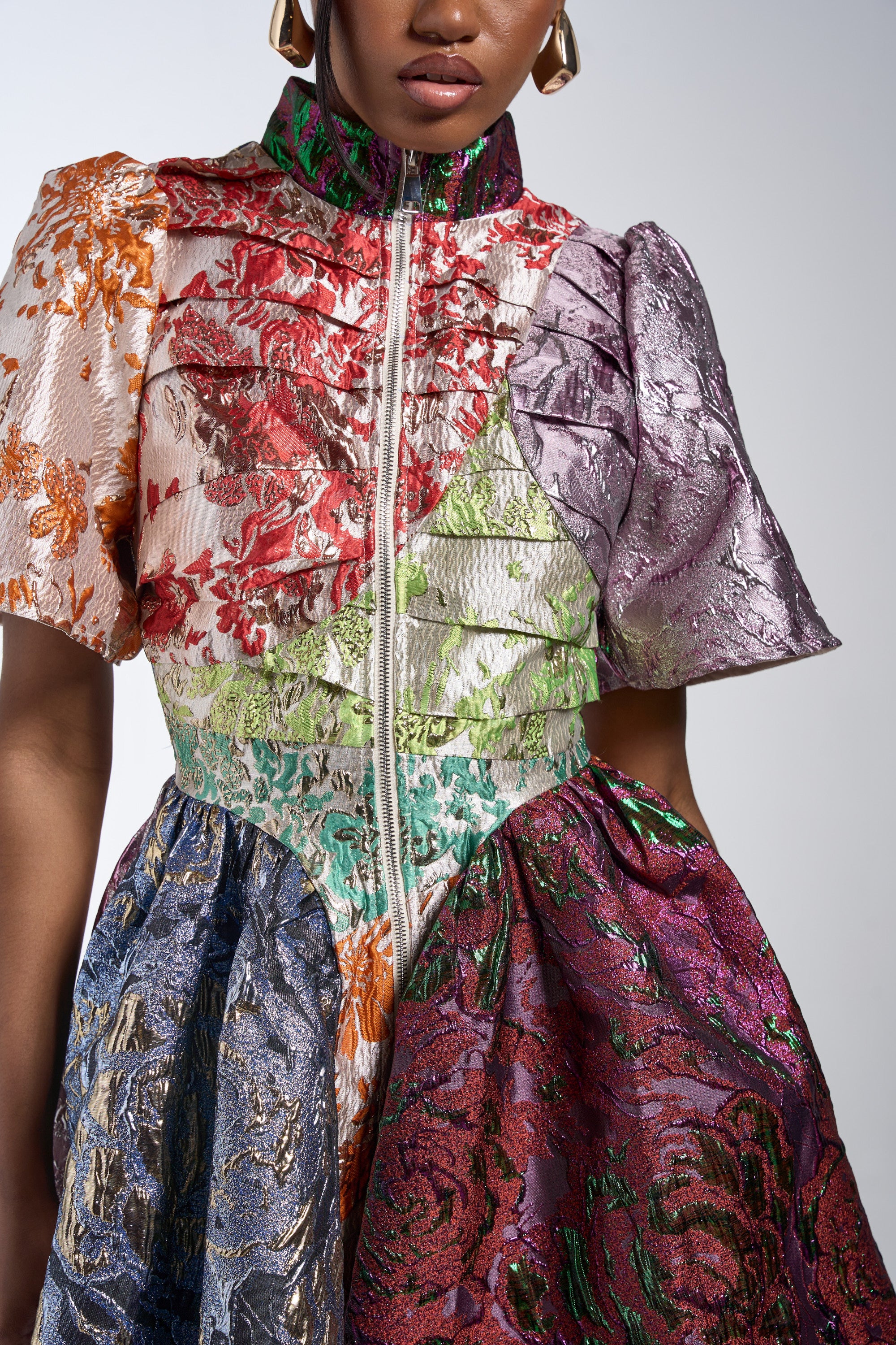 A person wears the STAY WITH ME BROCADE MINI DRESS, featuring a high collar, short sleeves, and metallic floral brocade in red, green, purple, blue, and gold. Only the upper body and part of the face are visible.