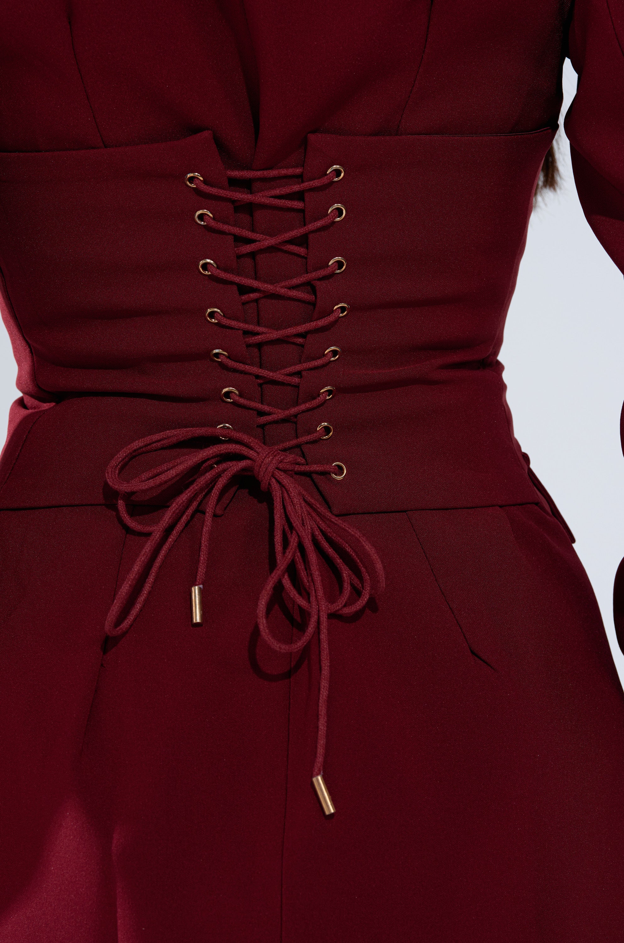 A close-up of a person wearing the BUSINESS CASUAL BLAZER DRESS IN BURGUNDY, featuring a corset-style lace-up front with gold eyelets and a neatly tied bow at the waist.