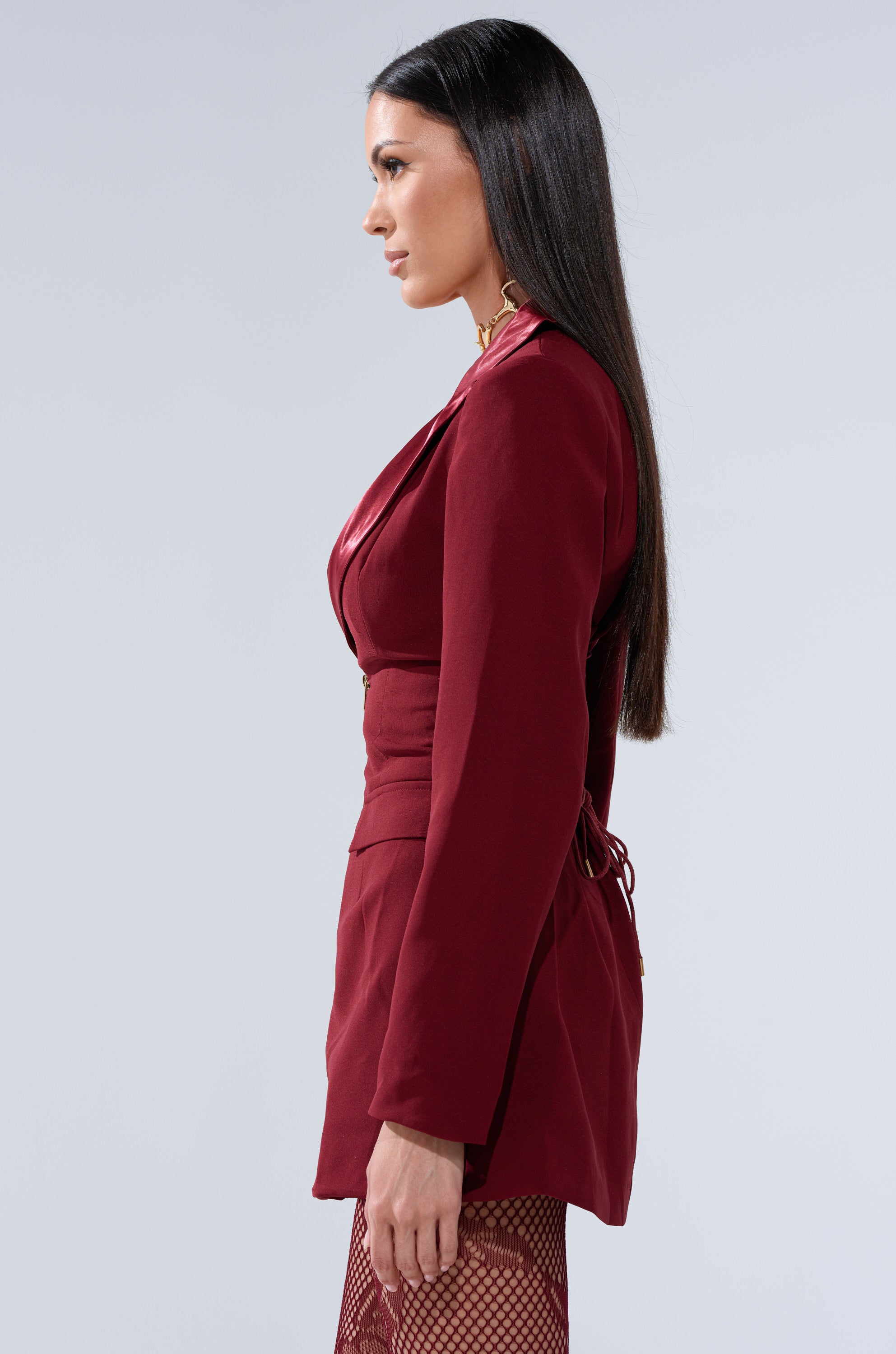 A woman with long straight dark hair wears the BUSINESS CASUAL BLAZER DRESS IN BURGUNDY and fishnet tights, standing in profile against a light gray background.