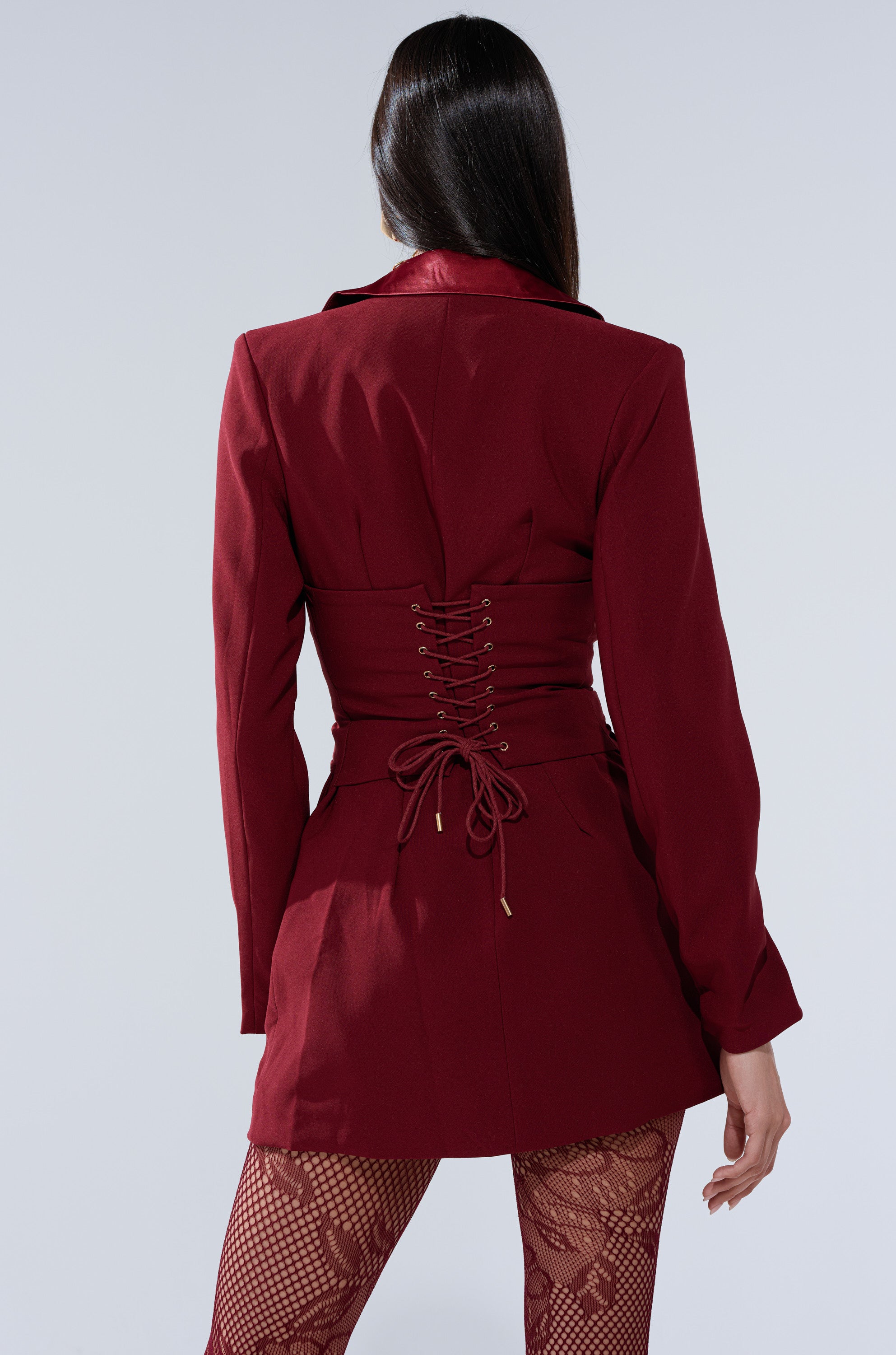 A woman with long dark hair models the BUSINESS CASUAL BLAZER DRESS IN BURGUNDY, featuring a lace-up corset back, styled with matching red fishnet tights, as she stands with her back to the camera against a light gray background.