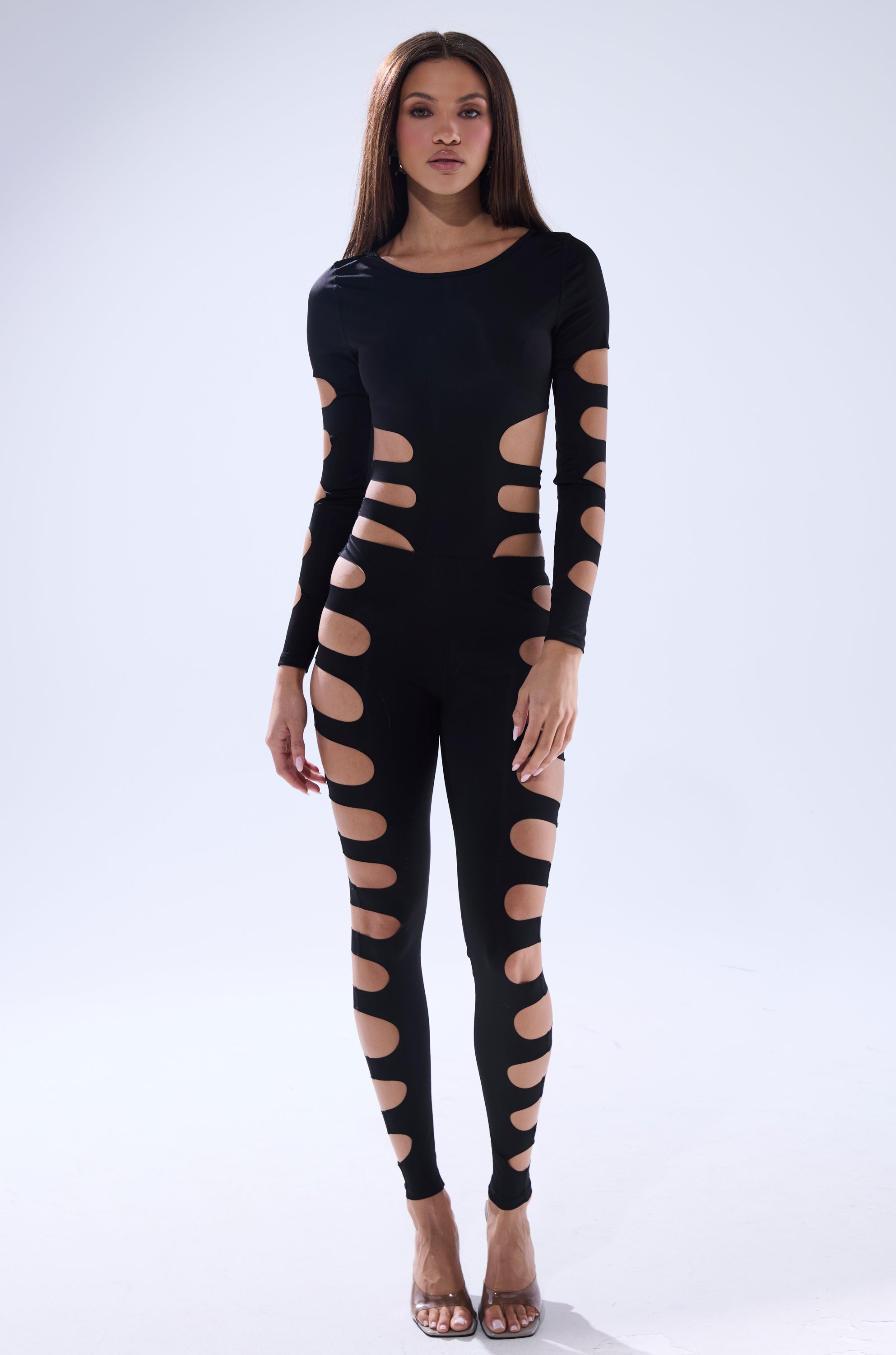 A woman with straight brown hair stands against a light background, wearing the OUT OF HIS LEAGUE JUMPSUIT featuring sleek style and multiple large cutout details on the arms, waist, and legs.